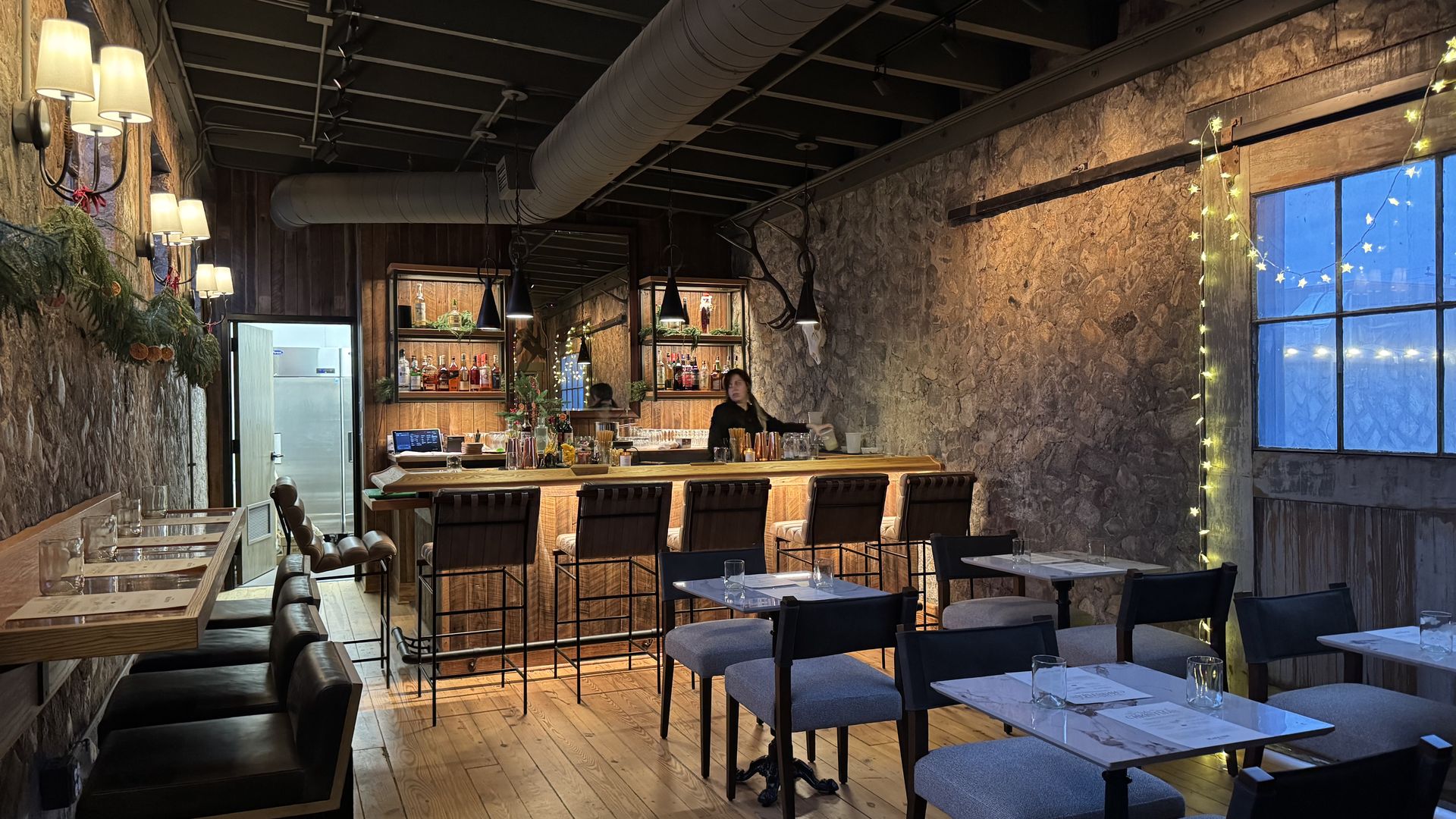 Cozy bar interior with stone walls, wooden floor, leather and fabric chairs, warm lighting, a bar counter with bottles, a person behind the bar, and string lights by a window.