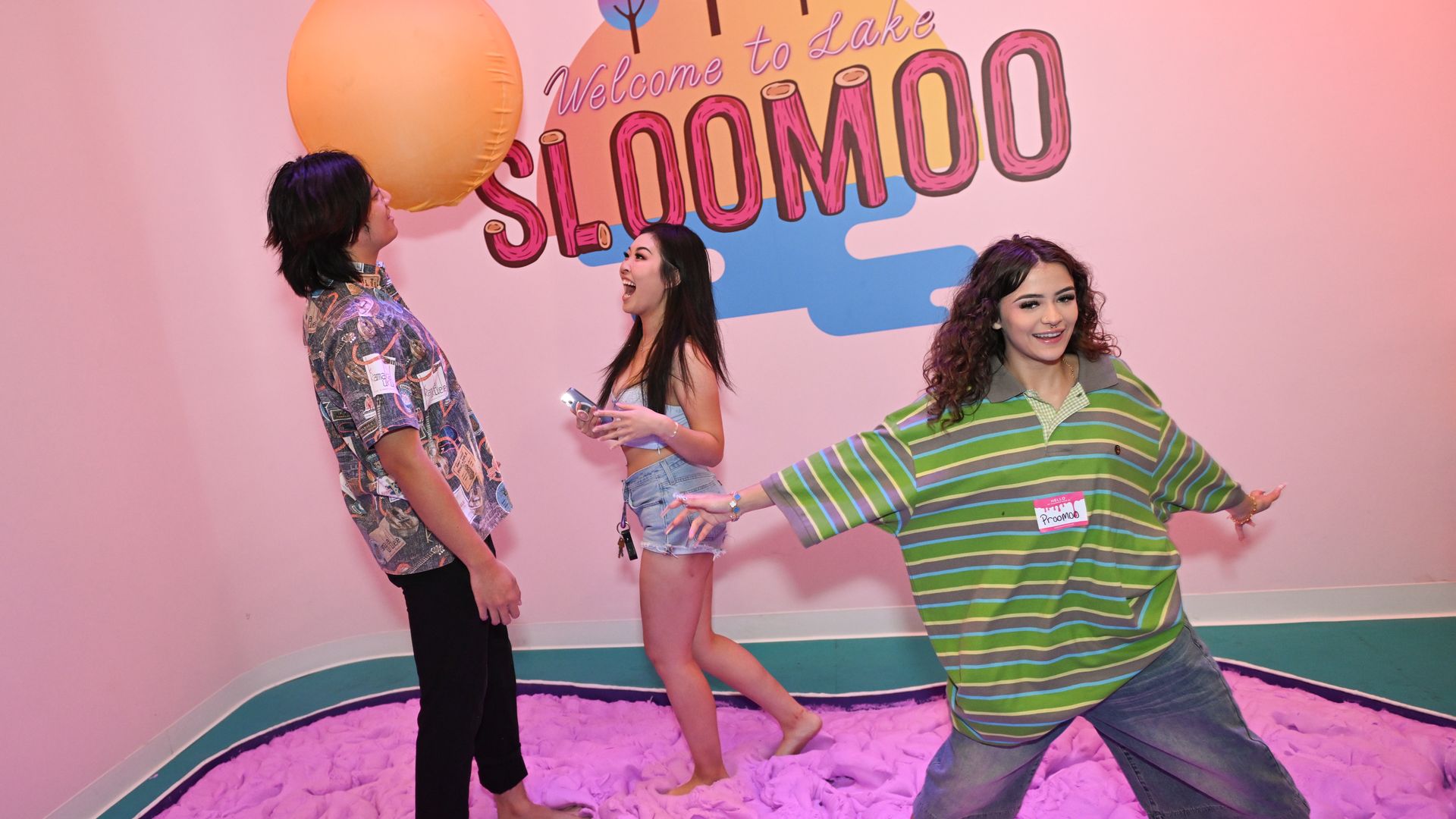 Three children pose in a pink photo booth with a mural reading "Welcome to Lake SLOOMO" in large pink letters at a Sloomoo Institute.