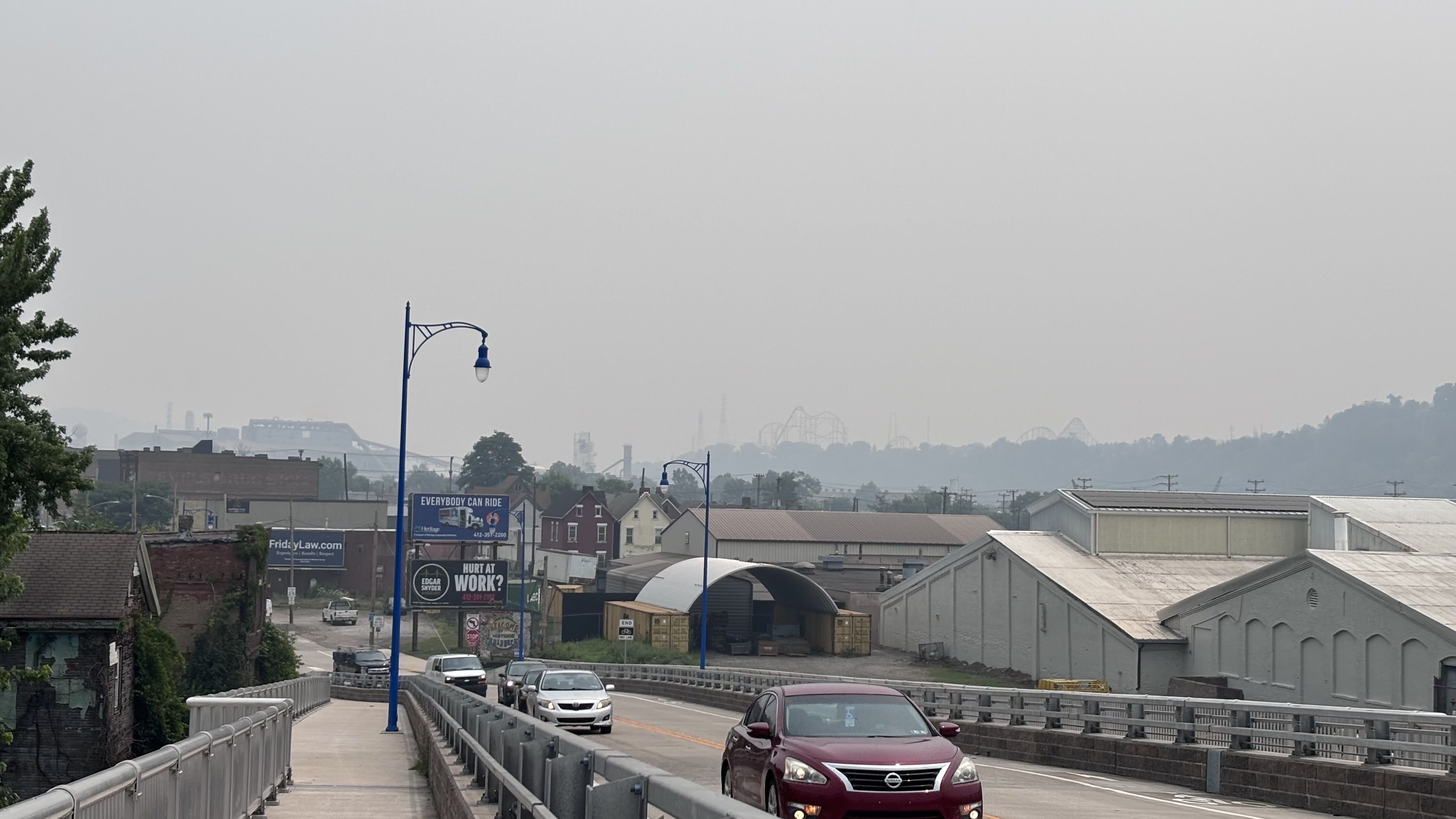 Wide city street with blue street lamps along a raised roadway, a red car in the foreground, hazy gray sky, and distant industrial buildings with roller coaster silhouettes on the horizon.