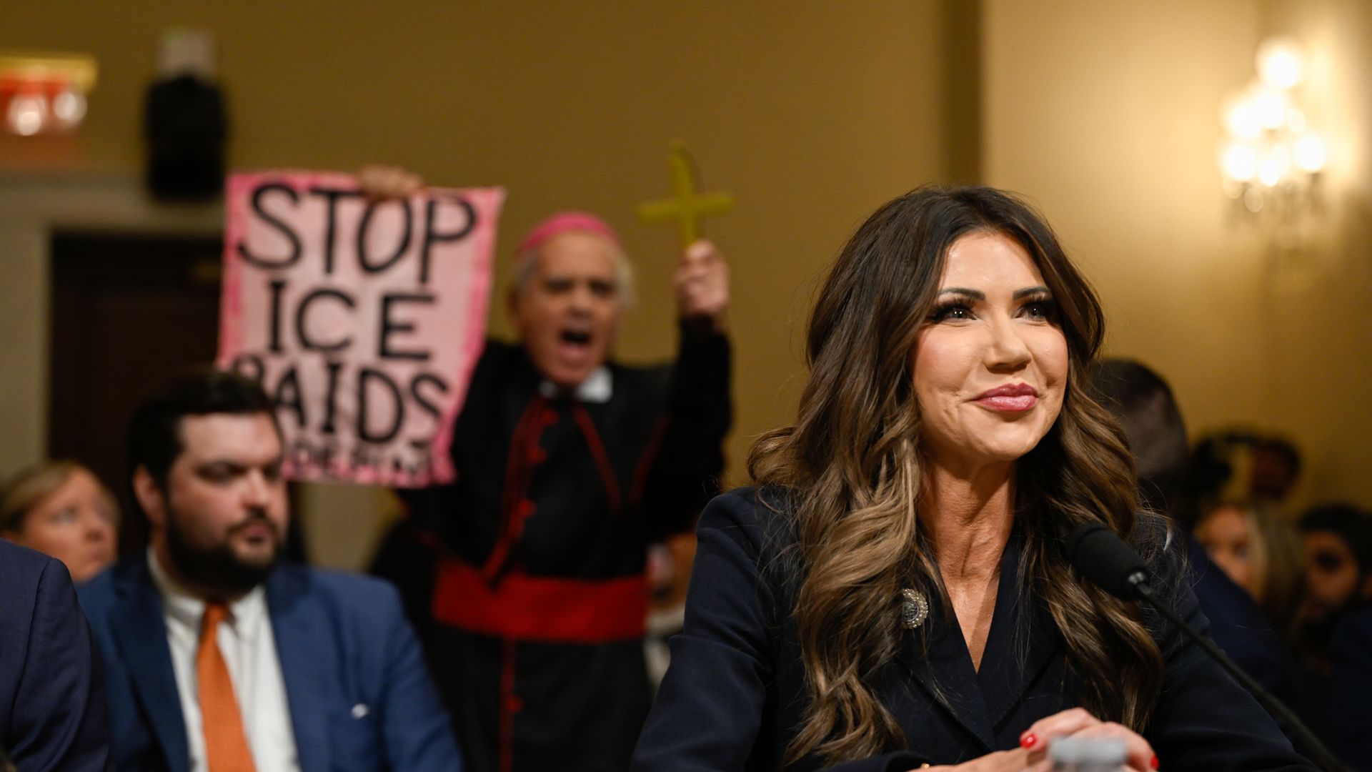 Kristi Noem smiles as she testifies to lawmakers (who are not pictured) while a man dressed as a clergy member holds a sign that says "STOP ICE RAIDS" and a cross behind her.