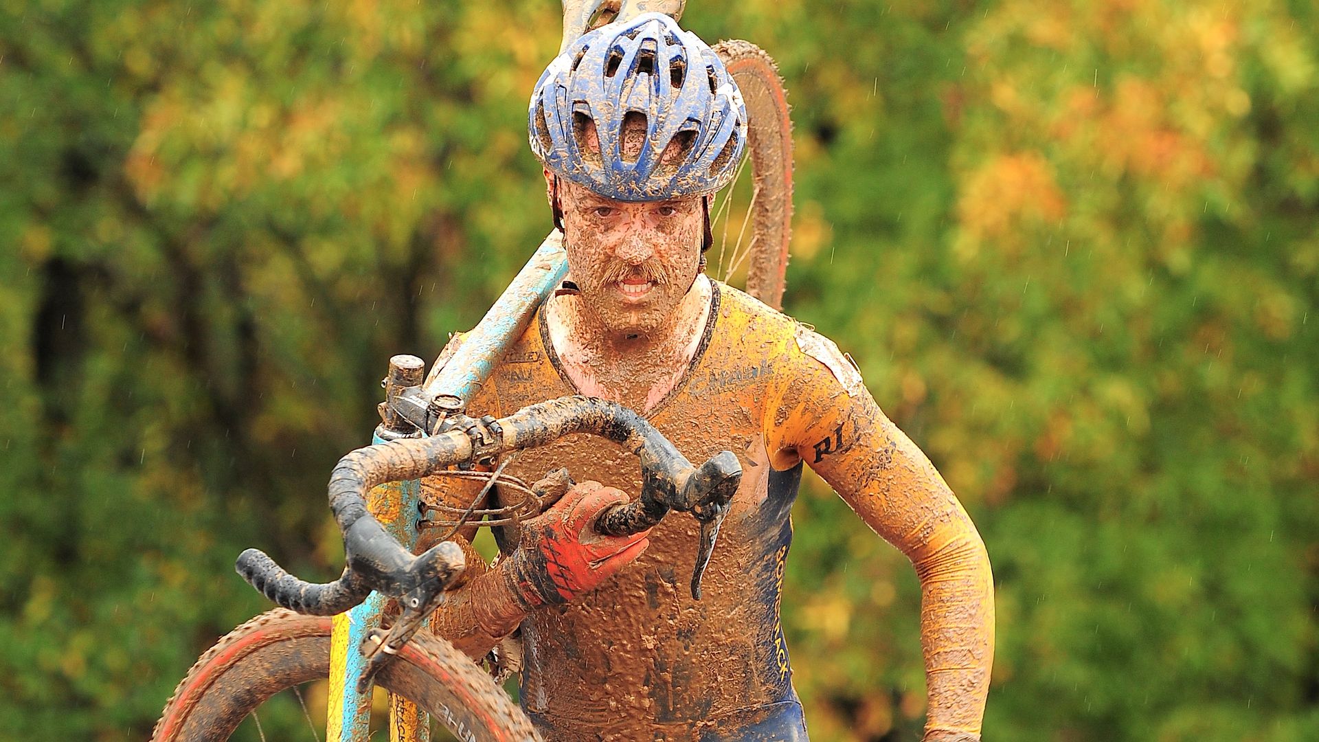 A cyclist covered in mud carries their bike on their shoulder while wearing a blue helmet and racing gear during a muddy cyclocross event. Trees with green and orange foliage are visible in the background.