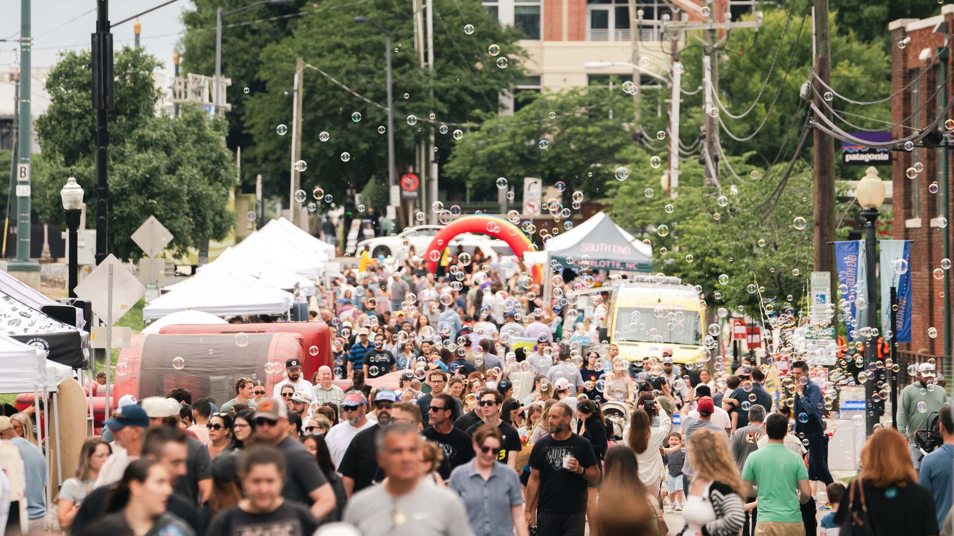 Camden Commons. Busy city street festival with many people walking among white tents, bubbles floating in the air, brick buildings, and green trees in the background.