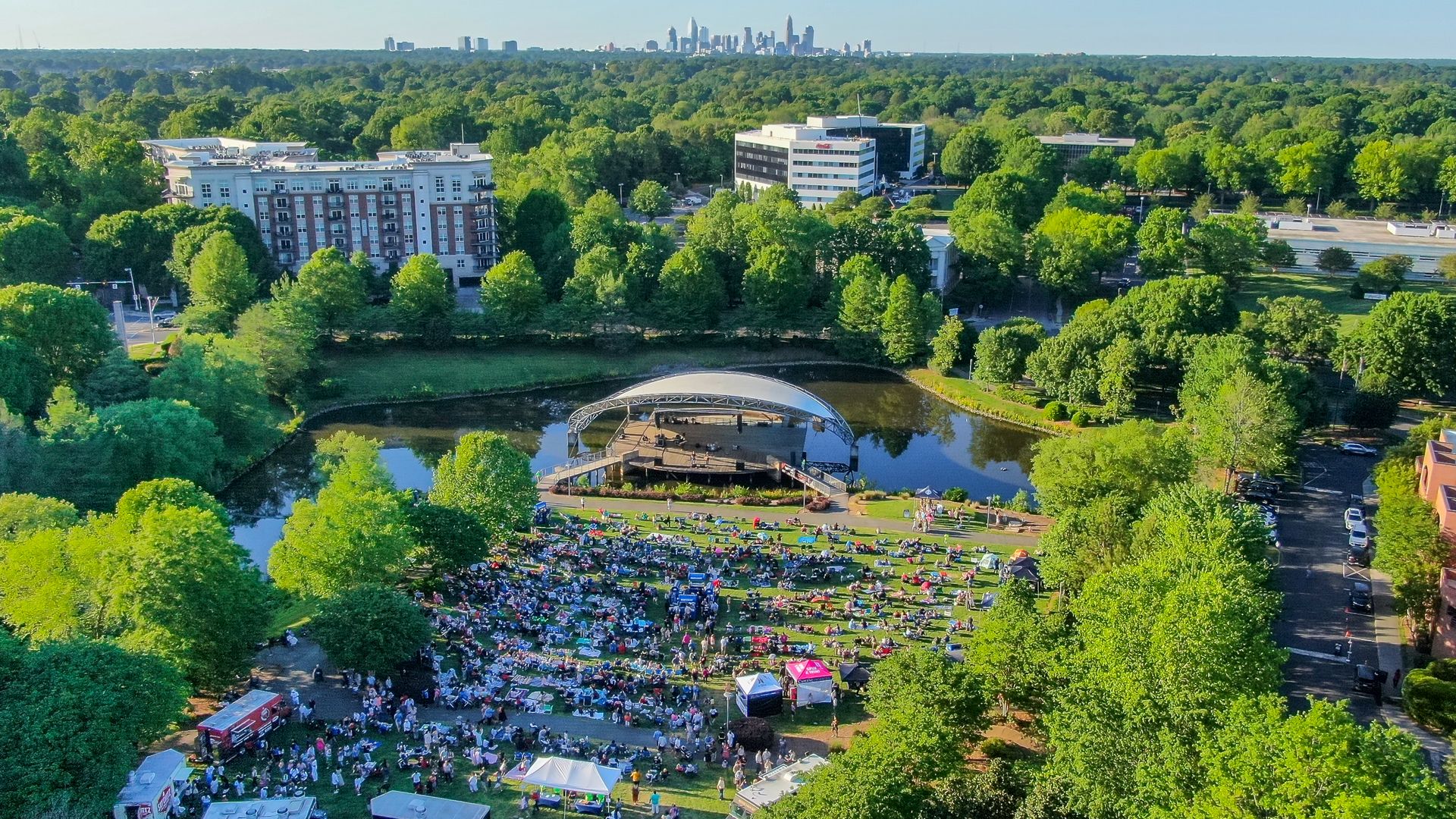Aerial view of a lush park with a large crowd on the lawn for an outdoor concert; a curved white canopy stage sits beside a small lake, with a distant city skyline under a clear blue sky.