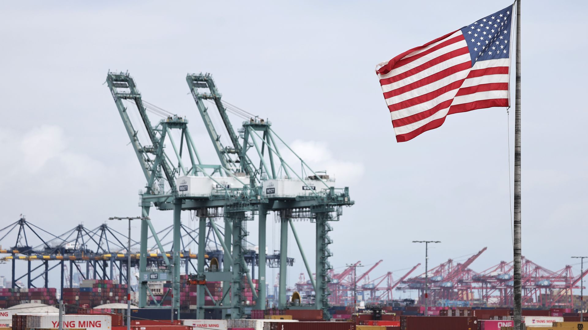 Shipping containers stacked up at a port, underneath giant cranes, with a US flag flapping in the breeze. 