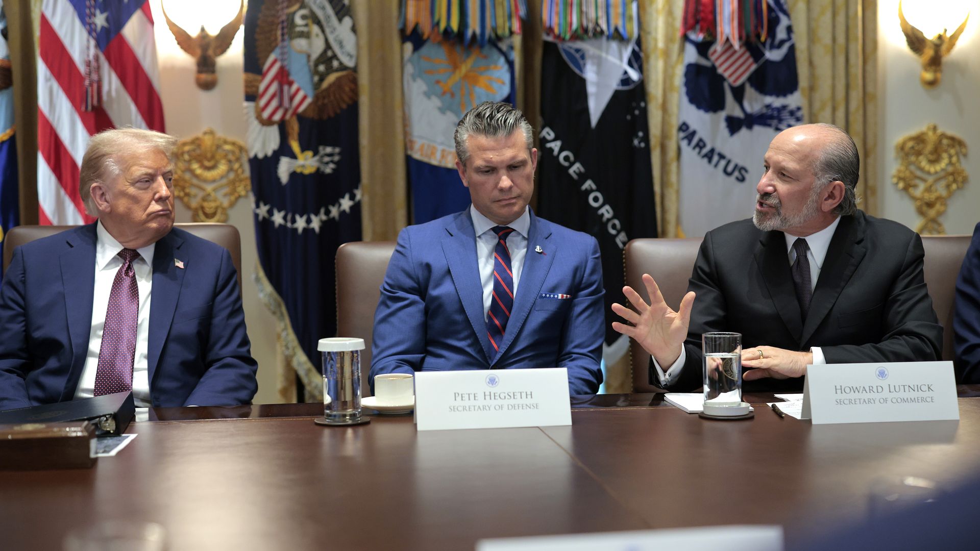 Three men, Donald Trump, Pete Hegseth and Howard Lutnick, are seated at a wooden table conversing. Military flags line the wall behind them. They are all in dark suits.