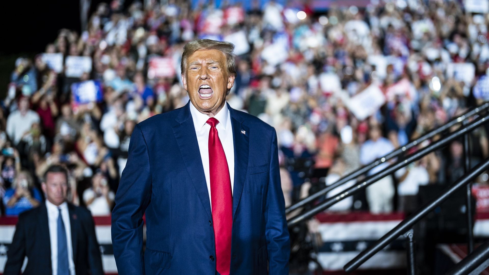 Former President Trump is shown in a blue suit, white shirt and red tie, standing in front of a crowd at a rally.