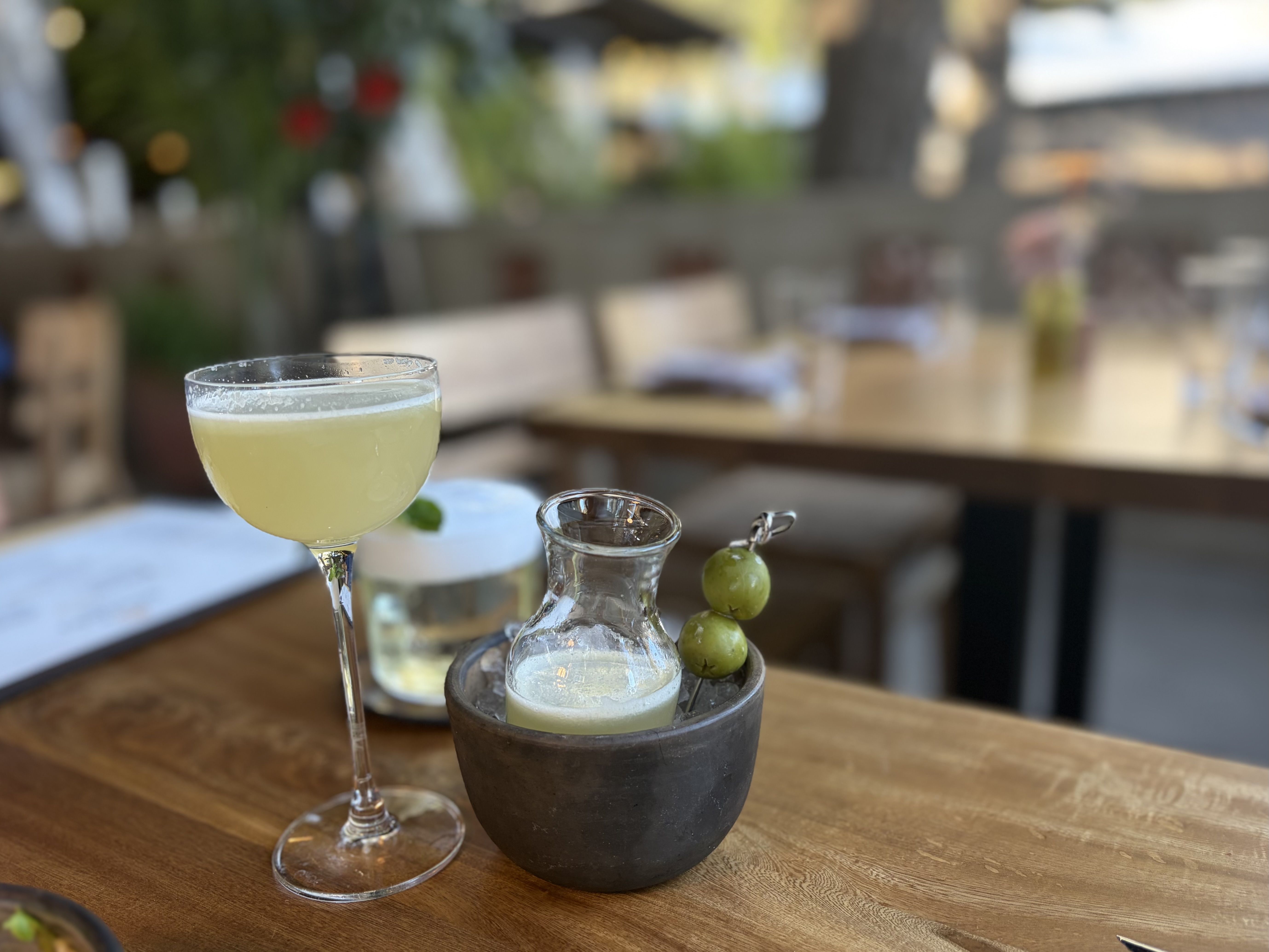 A pale yellow cocktail in a stemmed glass sits on a wooden table, with a water carafe and a grape skewer nearby; soft, blurred outdoor restaurant scene in the background.