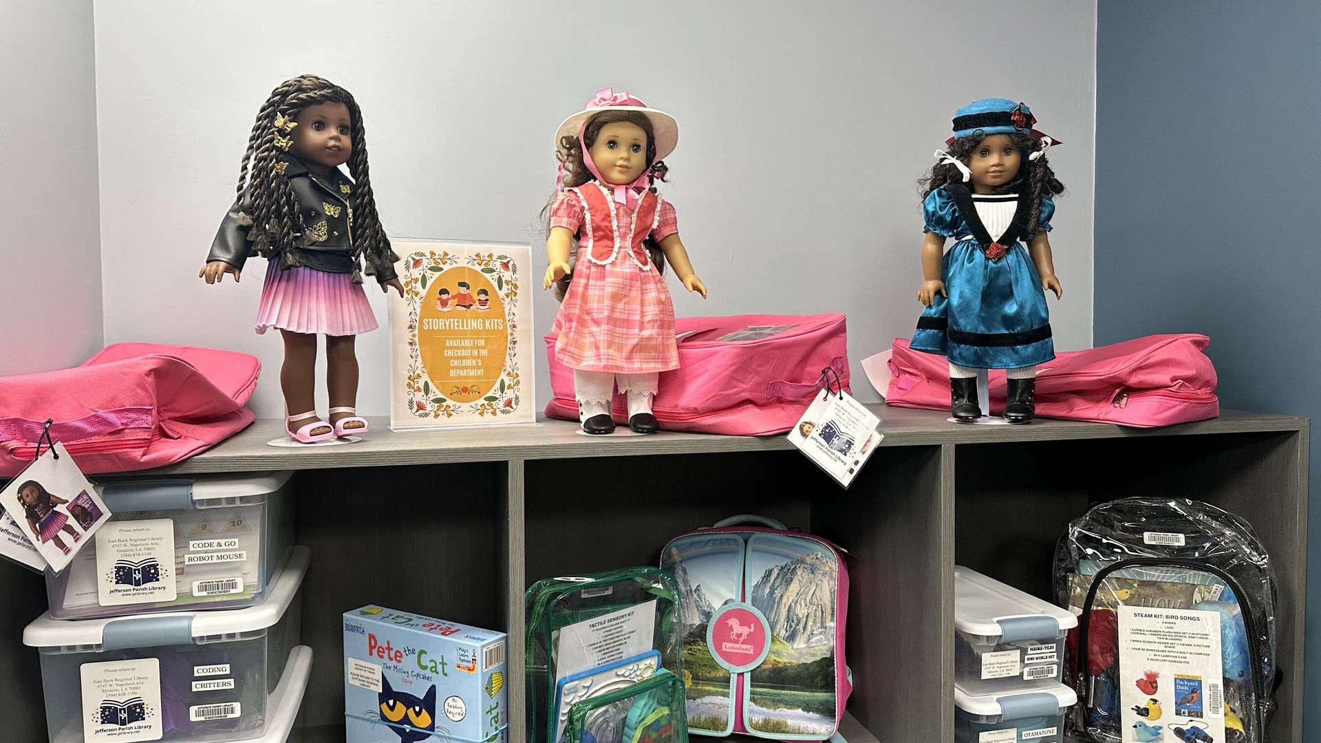 Three American Girl dolls on a shelf: left with braids and a black jacket, middle in pink plaid dress with sunhat, right in blue dress and hat. A "Storytelling Kits" sign and pink bags behind.