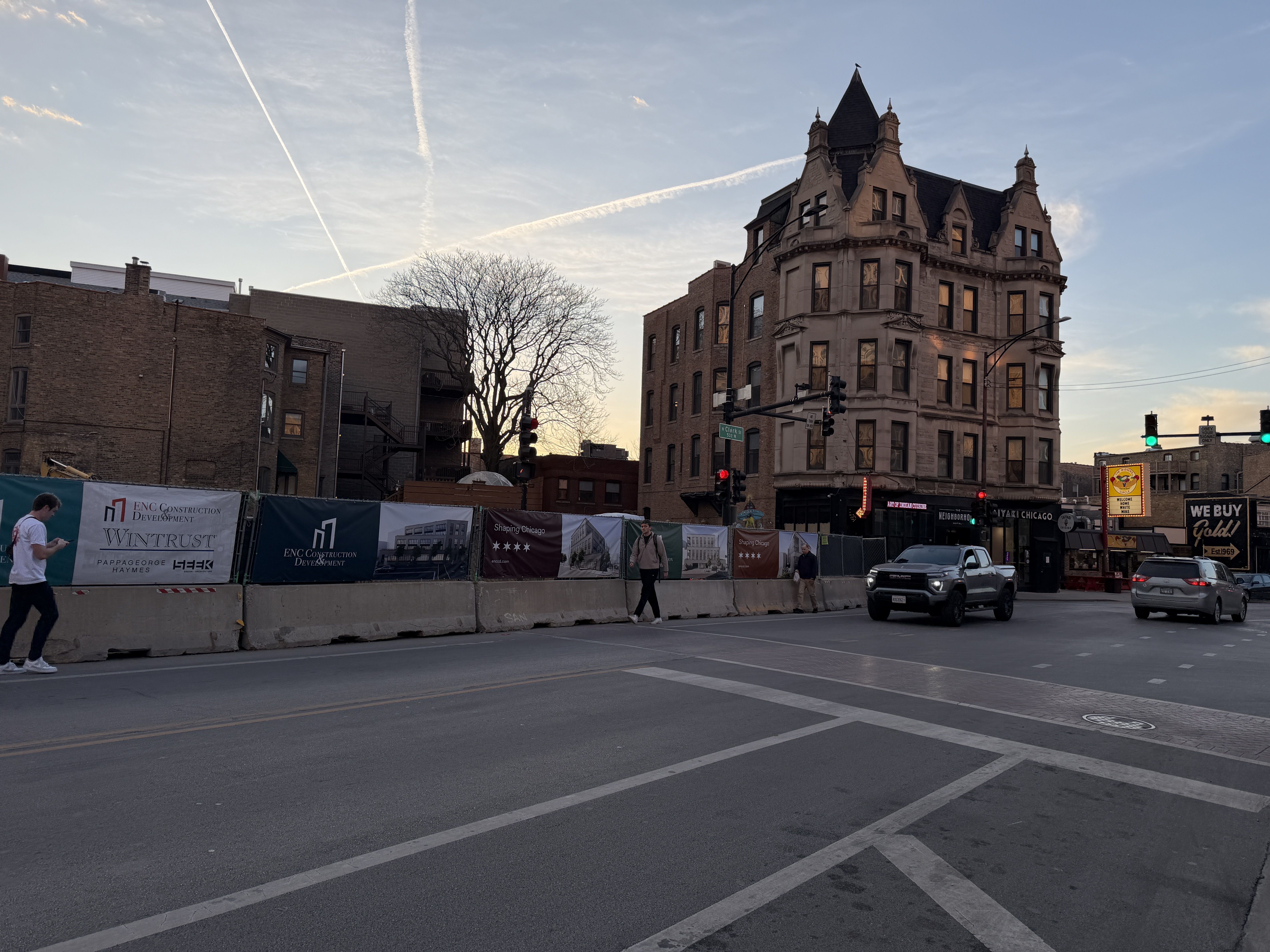 City intersection at dusk with a historic multi-story building, two vehicles on the road, construction barriers with banners, and people walking on the sidewalk under a sky with airplane trails.
