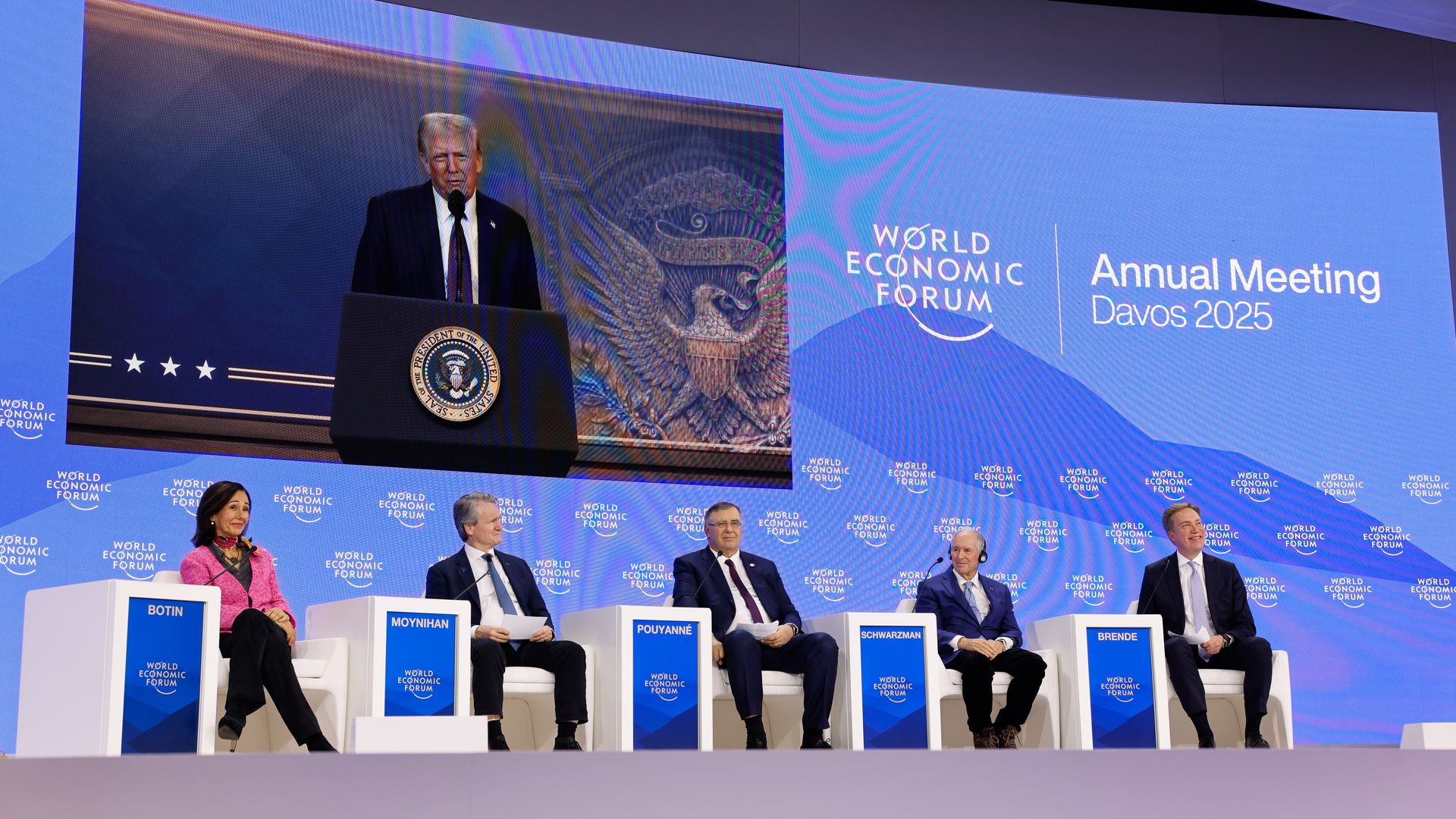 President Trump appears on a large screen as five people sit in chairs on a stage as he delivers a speech virtually at the World Economic Forum in Davos, Switzerland on Jan. 23, 2025.