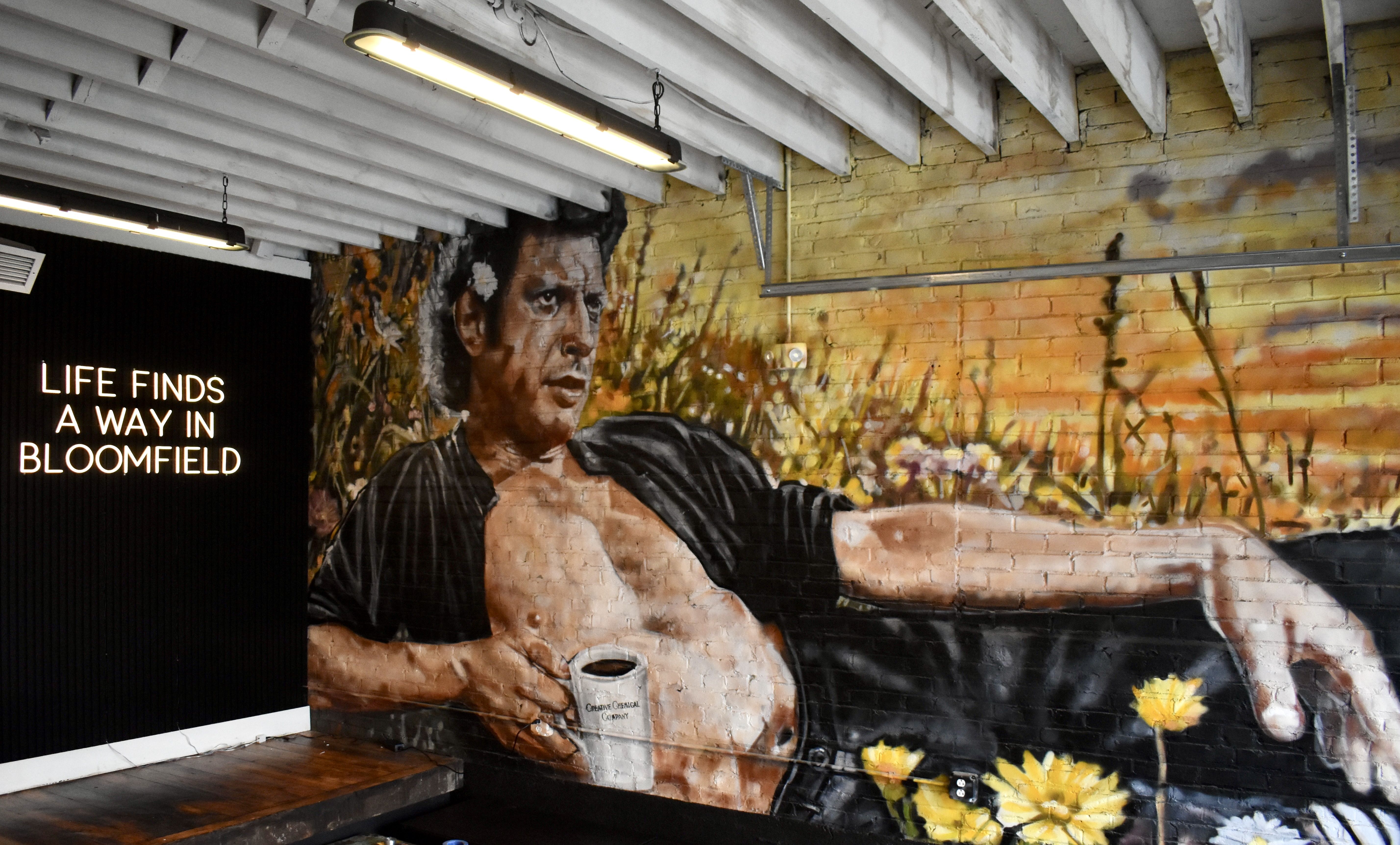 Mural of a shirtless man reclining with coffee mug labeled "Orange Chemical Company," surrounded by yellow flowers on a brick wall with neon text "Life finds a way in Bloomfield."