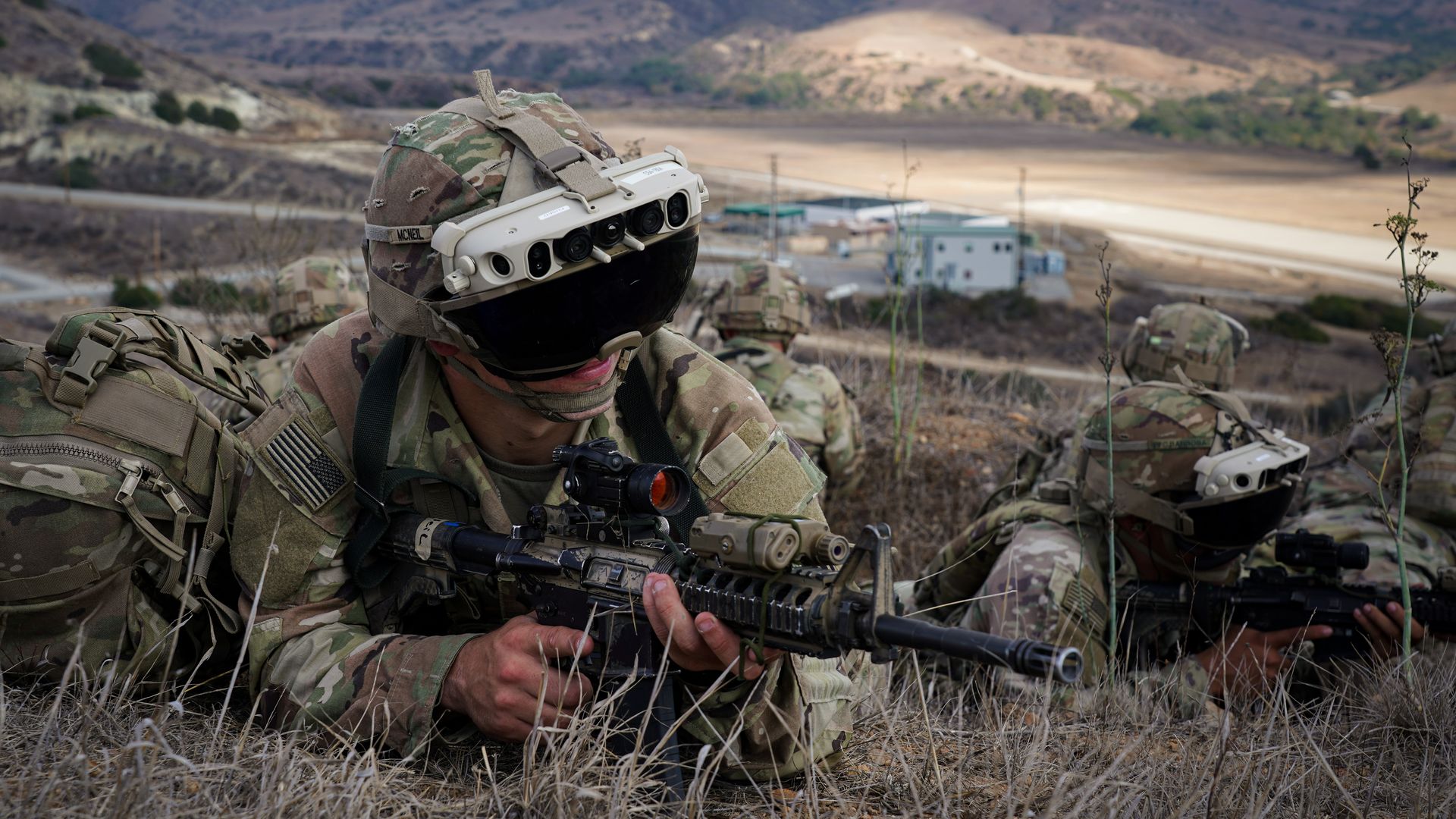 U.S. soldiers are prone in a field. At least two of them are wearing the Army's futuristic goggles, the IVAS. Mountains are seen in the background.