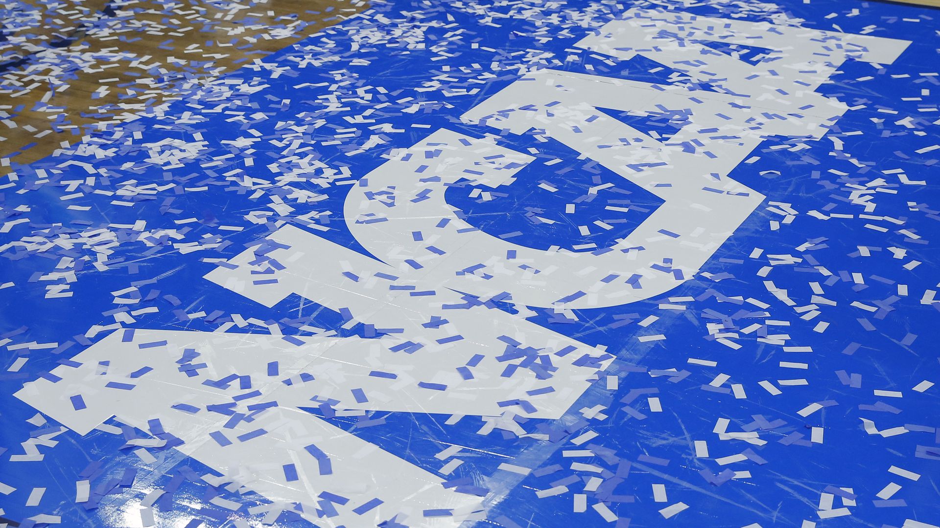 In this image, the blue and white block letter logo for NCAA is on the floor of a basketball court as confetti falls onto the floor.