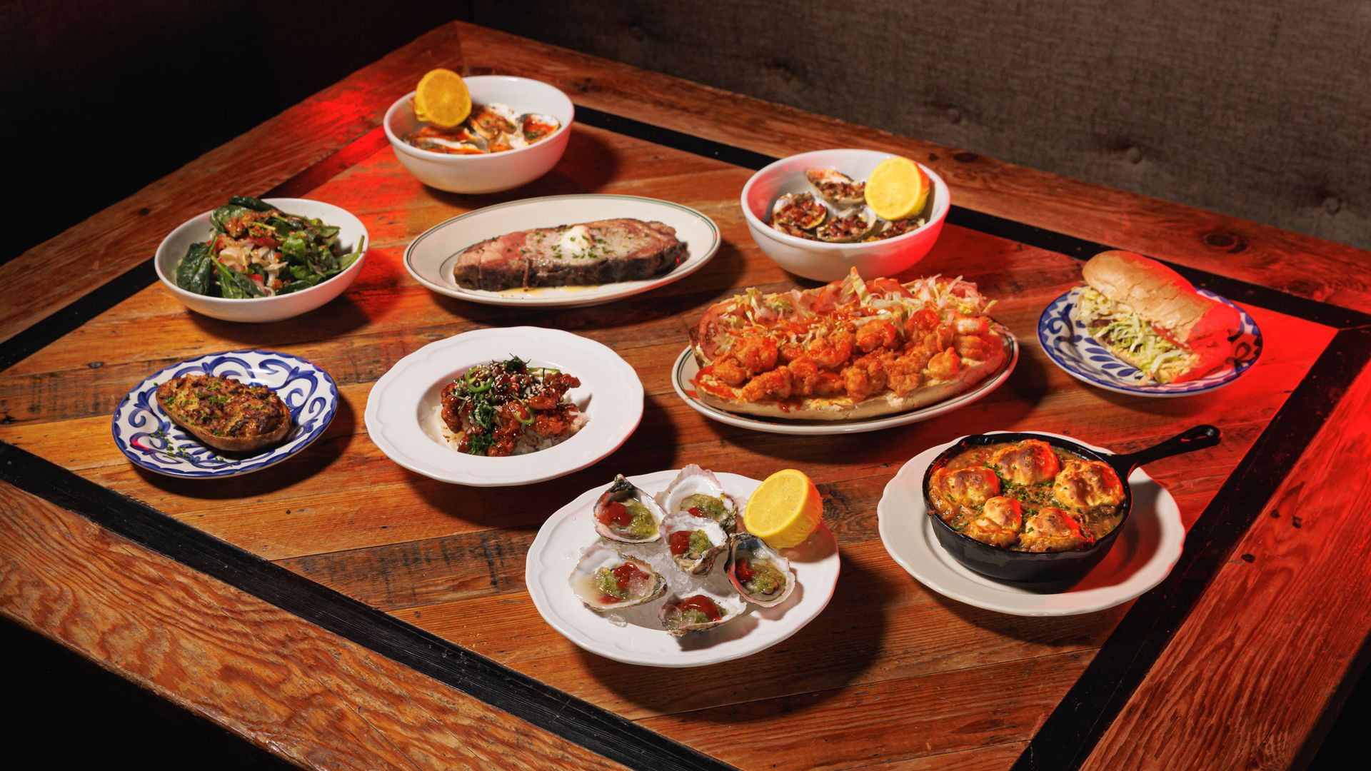 Spread of food on a wooden table: slices of steak, bowls of seafood with lemon wedges, a platter of fried bites, a sandwich with shredded lettuce, and a greens salad under warm red lighting.