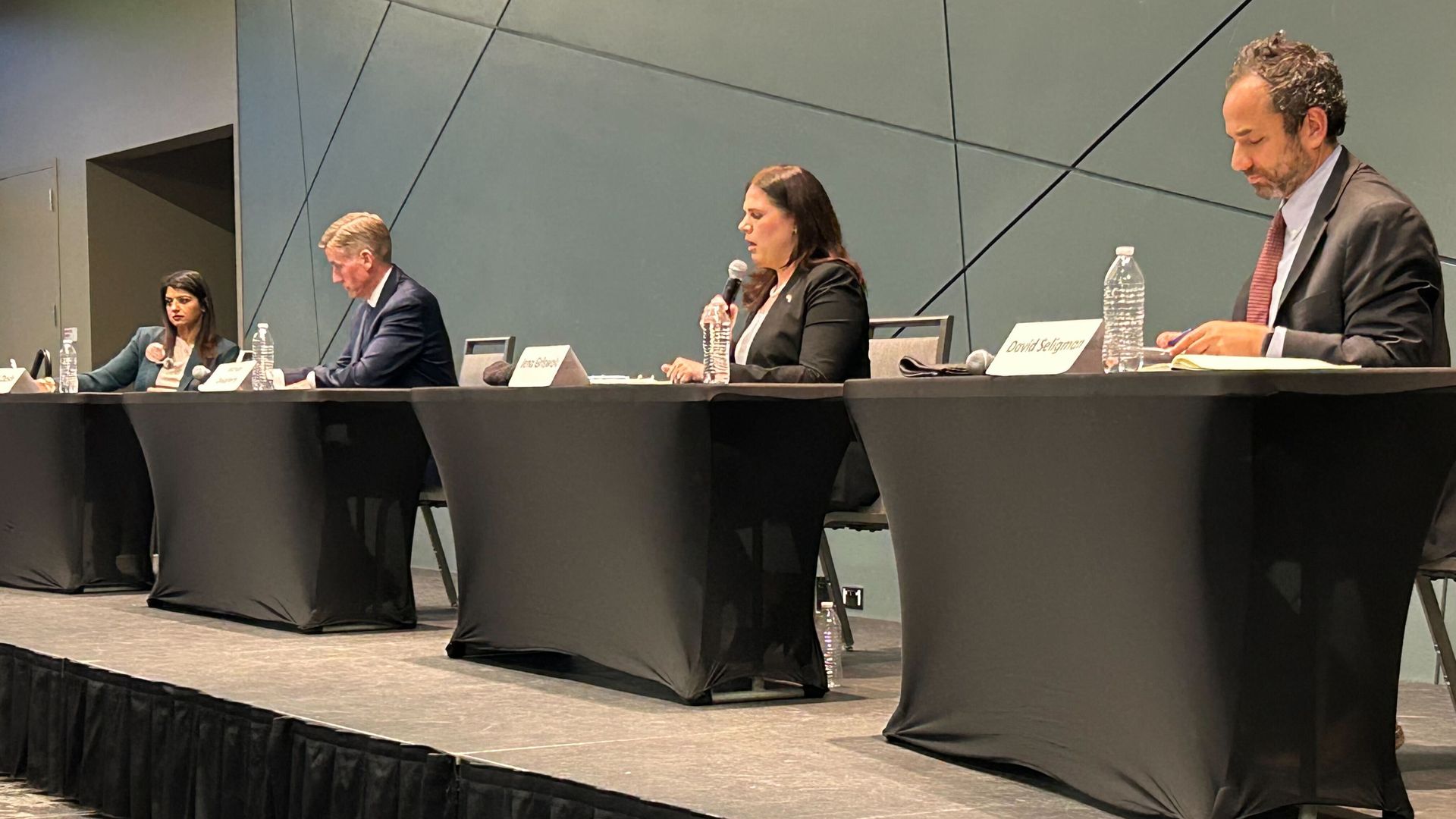 Panel of four professionals at a long black-draped table on a stage; two women and two men, microphones and water bottles in front; nameplates visible; geometric wall panels behind.