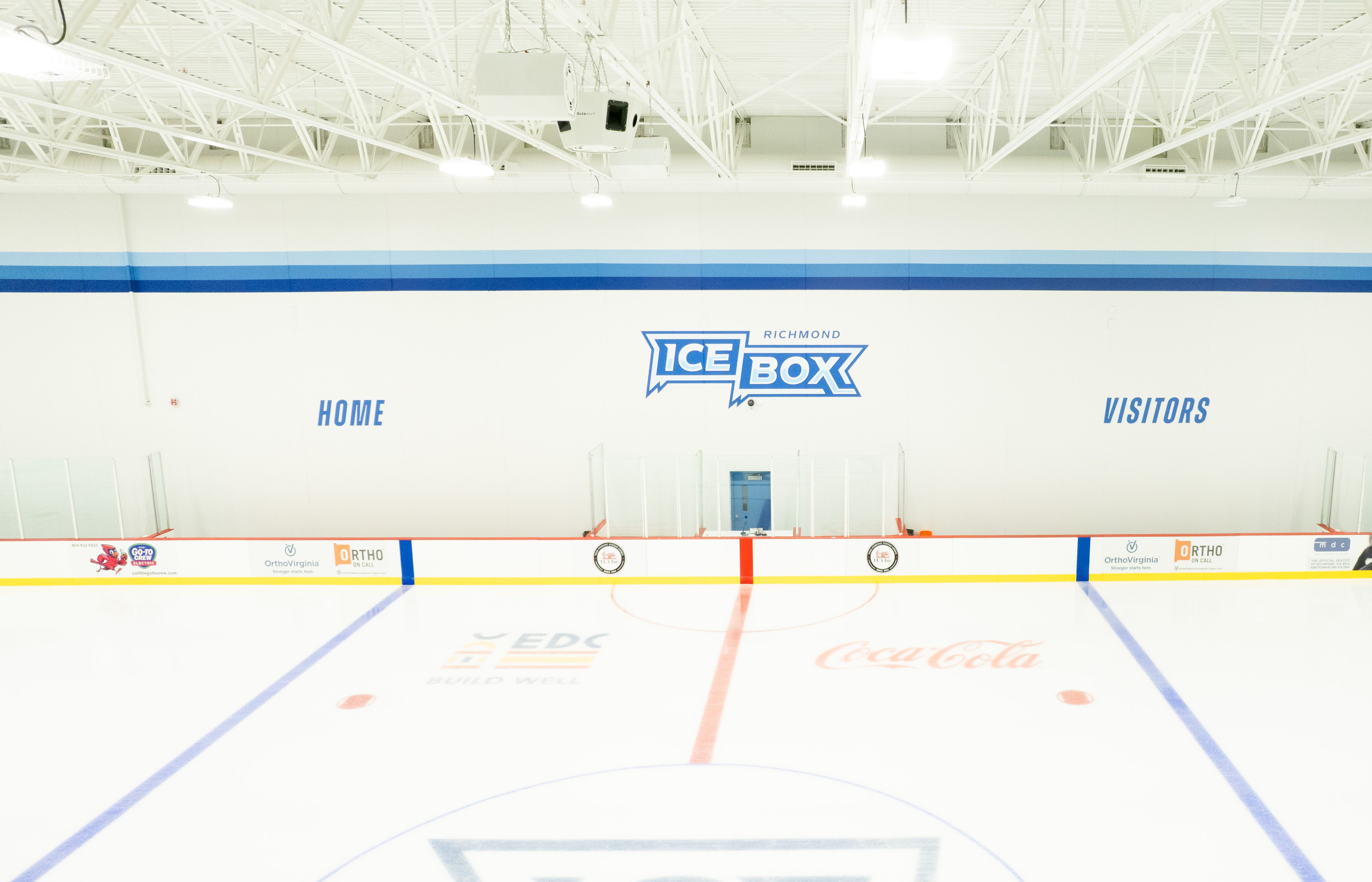 Empty indoor ice rink with white walls, blue stripes, and a large "ICE BOX" Richmond logo. "HOME" and "VISITORS" signs on boards; glass, bright lights, and exposed metal trusses overhead.