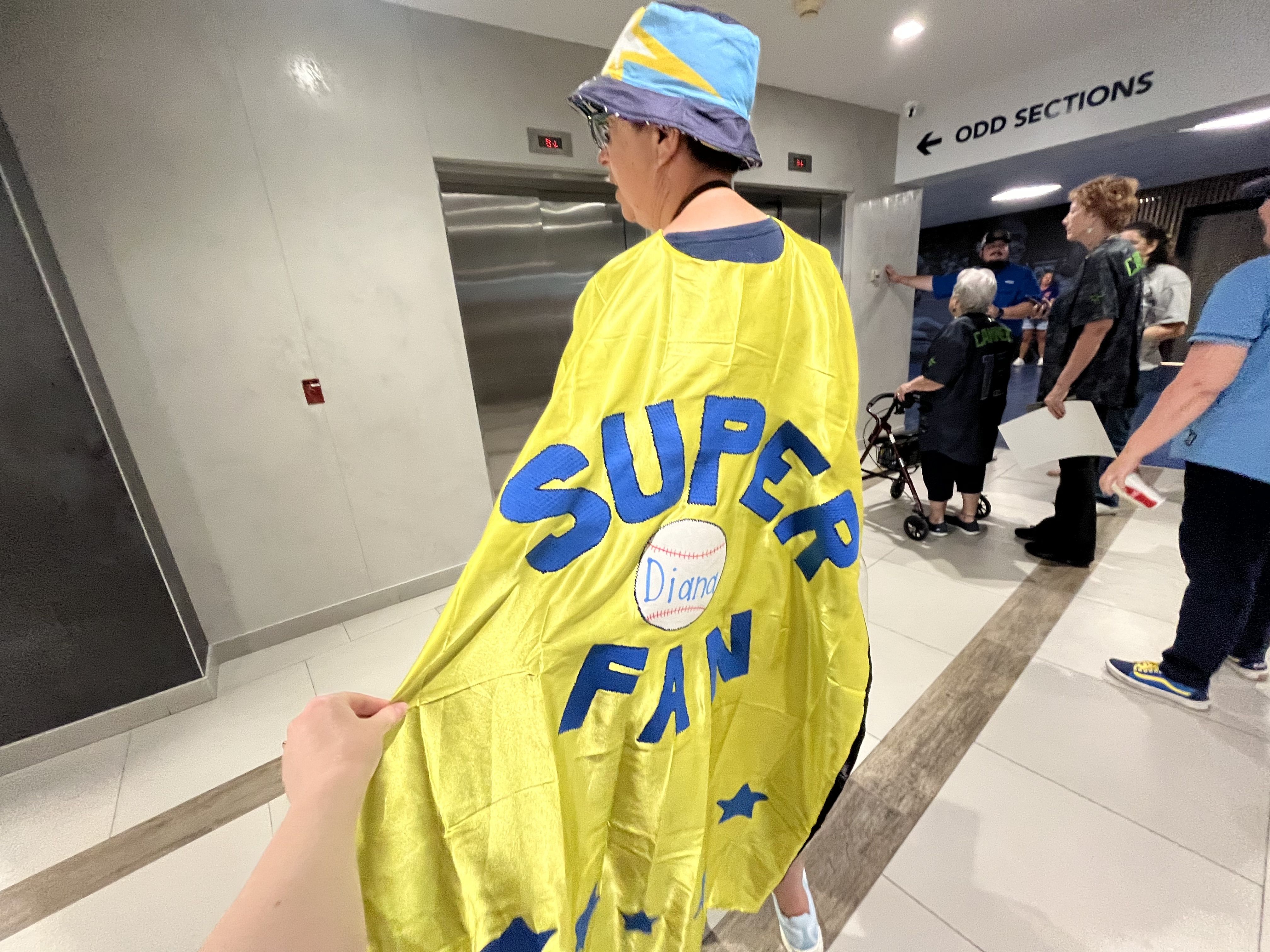 Back view of a person in a bright yellow cape that reads "SUPER FAN" with a baseball patch; a busy indoor hallway with others and an sign reading "ODD SECTIONS" in the background.