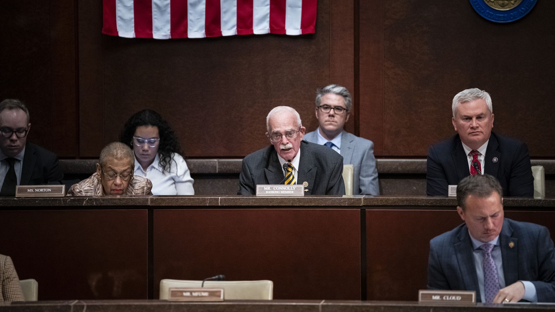 Rep. Gerry Connolly, wearing a gray suit and speaking at a wooden dais while flanked by colleagues.