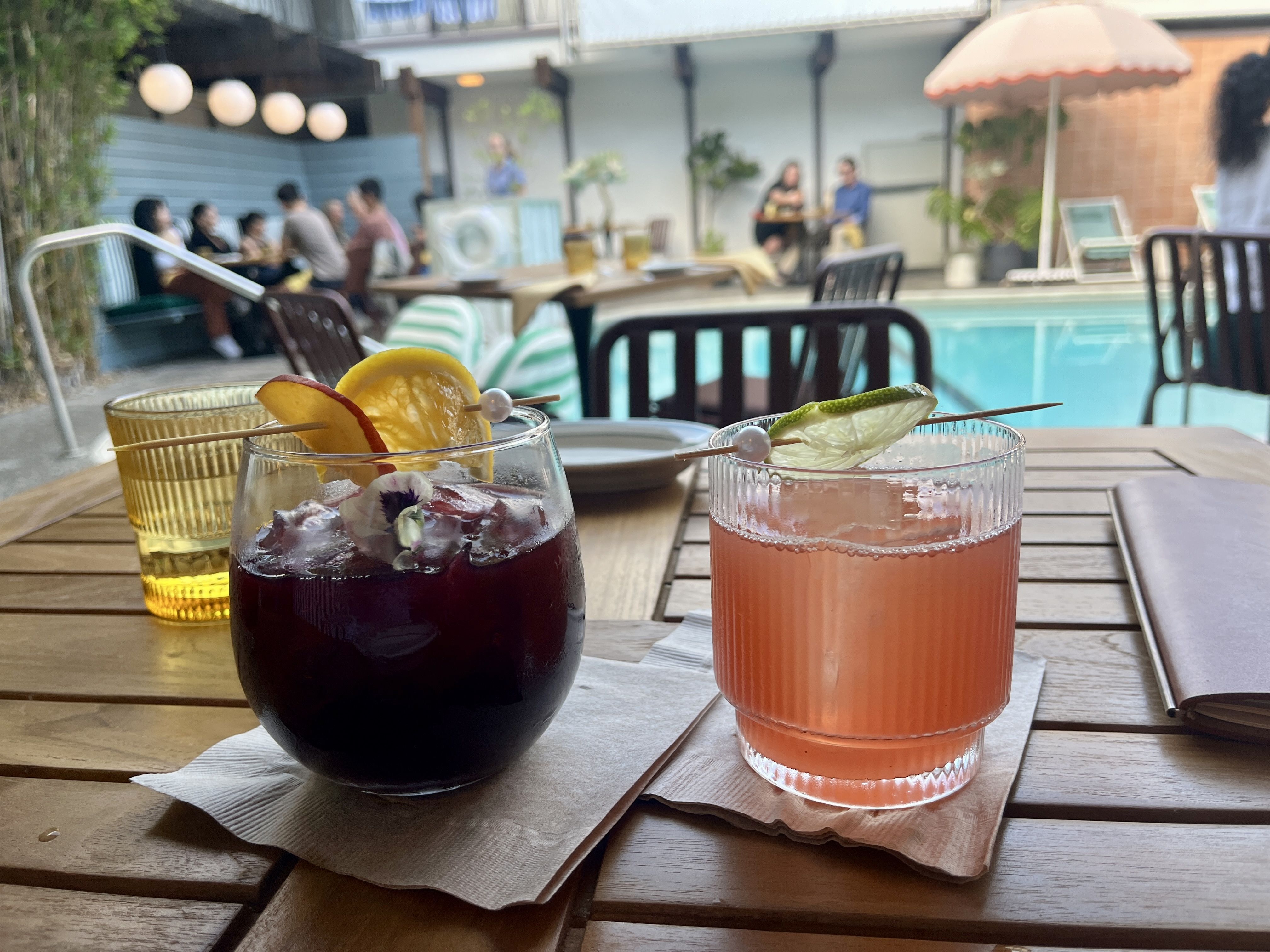 A glass of red sangria and a pink, tequila cocktail on a table with a pool in the background.