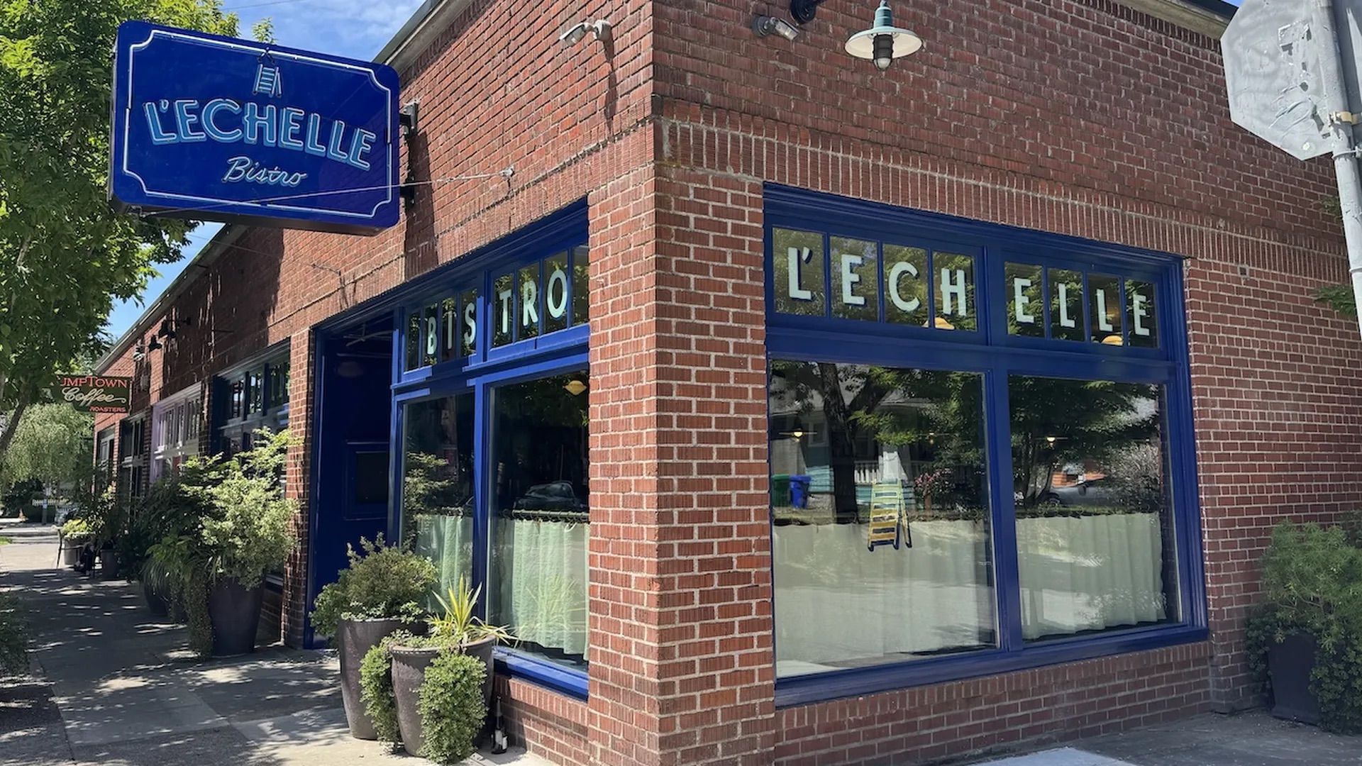 Corner of a red brick building with blue trim and a large blue sign reading "L'ÉCHELLE Bistro." Wide windows reflect trees; potted plants line the sidewalk on a sunny day.