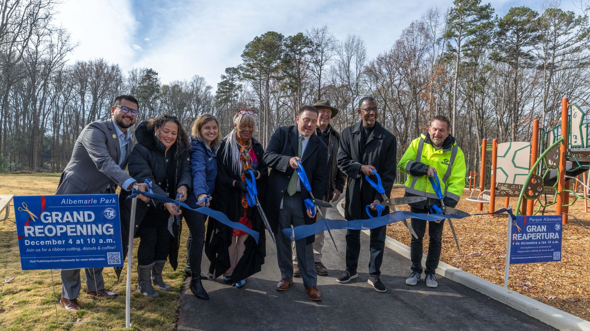 Eight people cutting a blue ribbon at Albemarle Park's grand reopening, with signs announcing December 4 at 10 a.m. The group holds large blue scissors near a playground and wooded area.