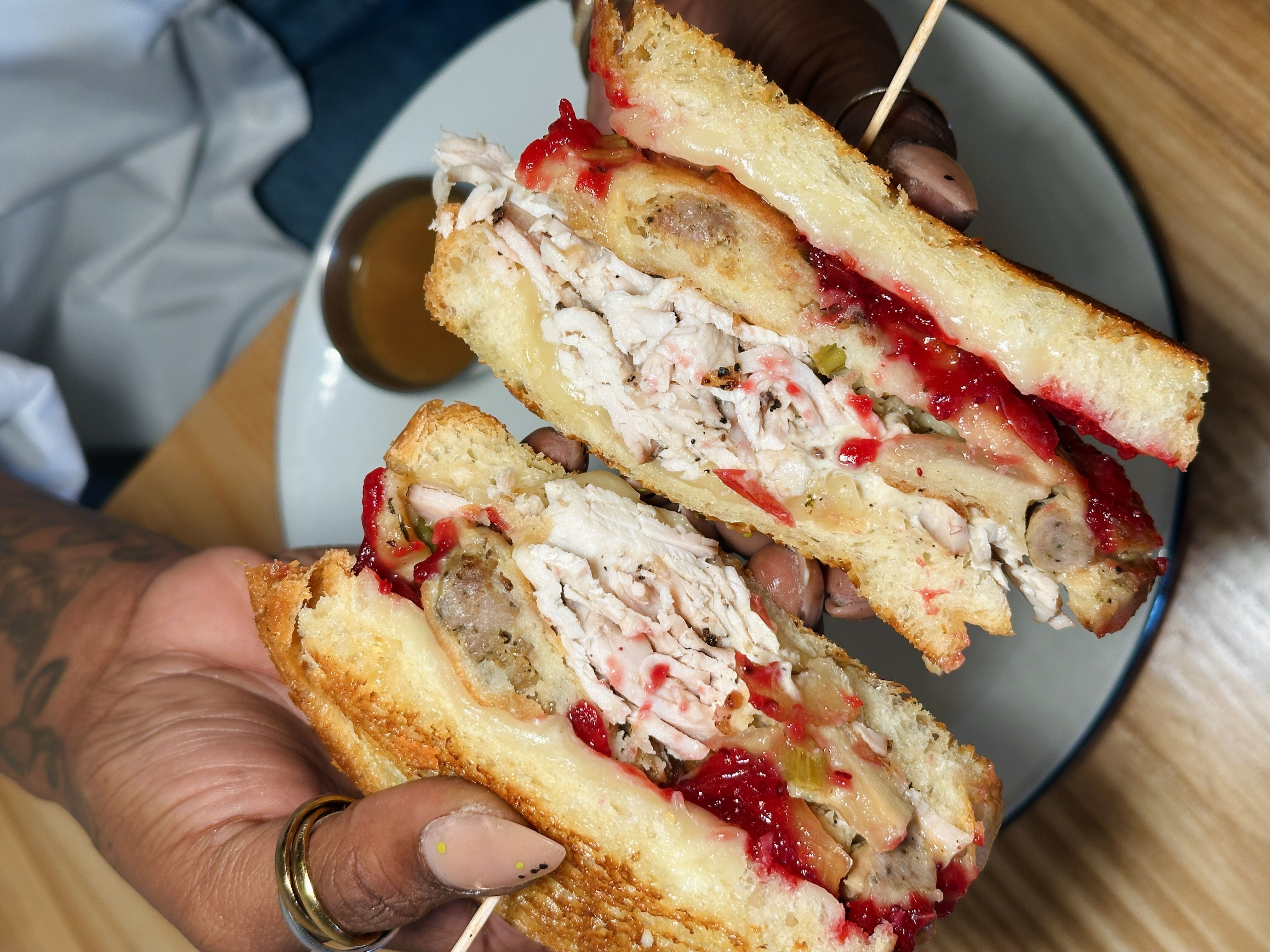 Hands holding a sandwich with white bread, sliced turkey, sausage, melted cheese, and red cranberry sauce, with a plate and brown gravy in the background.