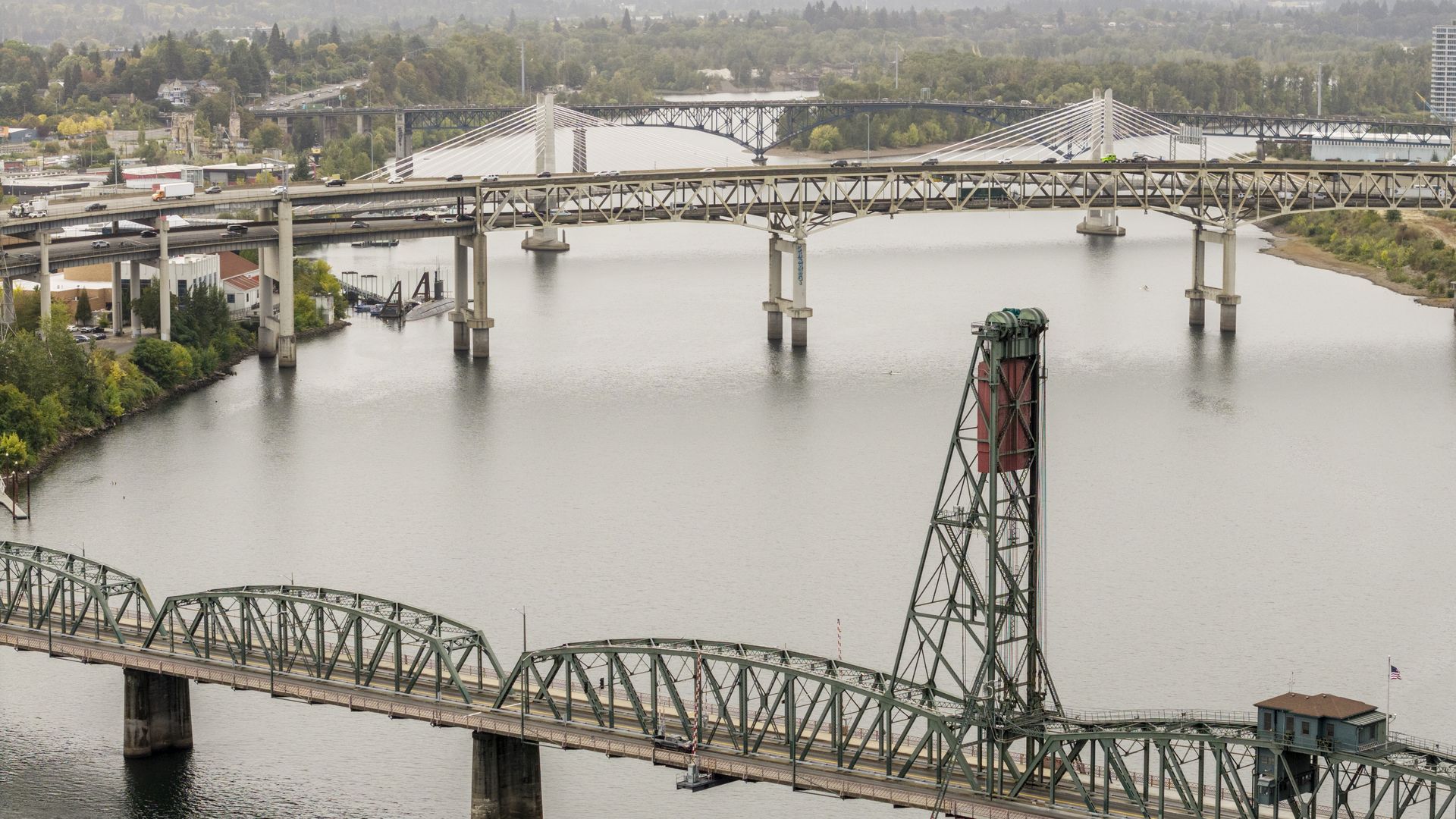 Wide river with multiple bridges: a large green truss lift bridge in the foreground, a white cable-stayed bridge behind, and a distant tree-lined cityscape.