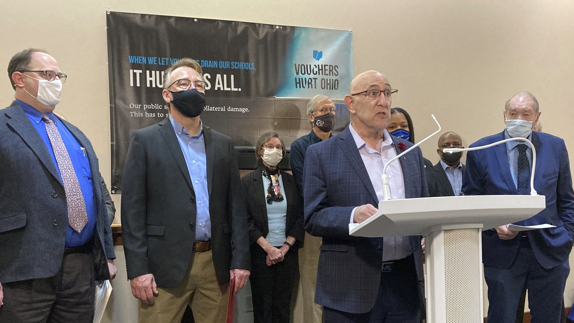 Columbus school board member Eric Brown stands at a podium with a group of masked people and a "Vouchers Hurt Ohio" banner behind him
