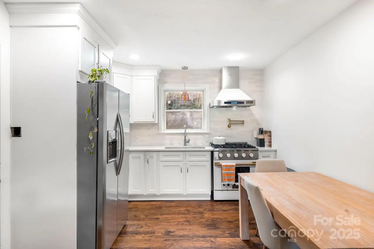 Bright modern kitchen with white cabinets, stainless steel refrigerator and stove, wood floor, small window above sink, wooden dining table with two beige chairs.