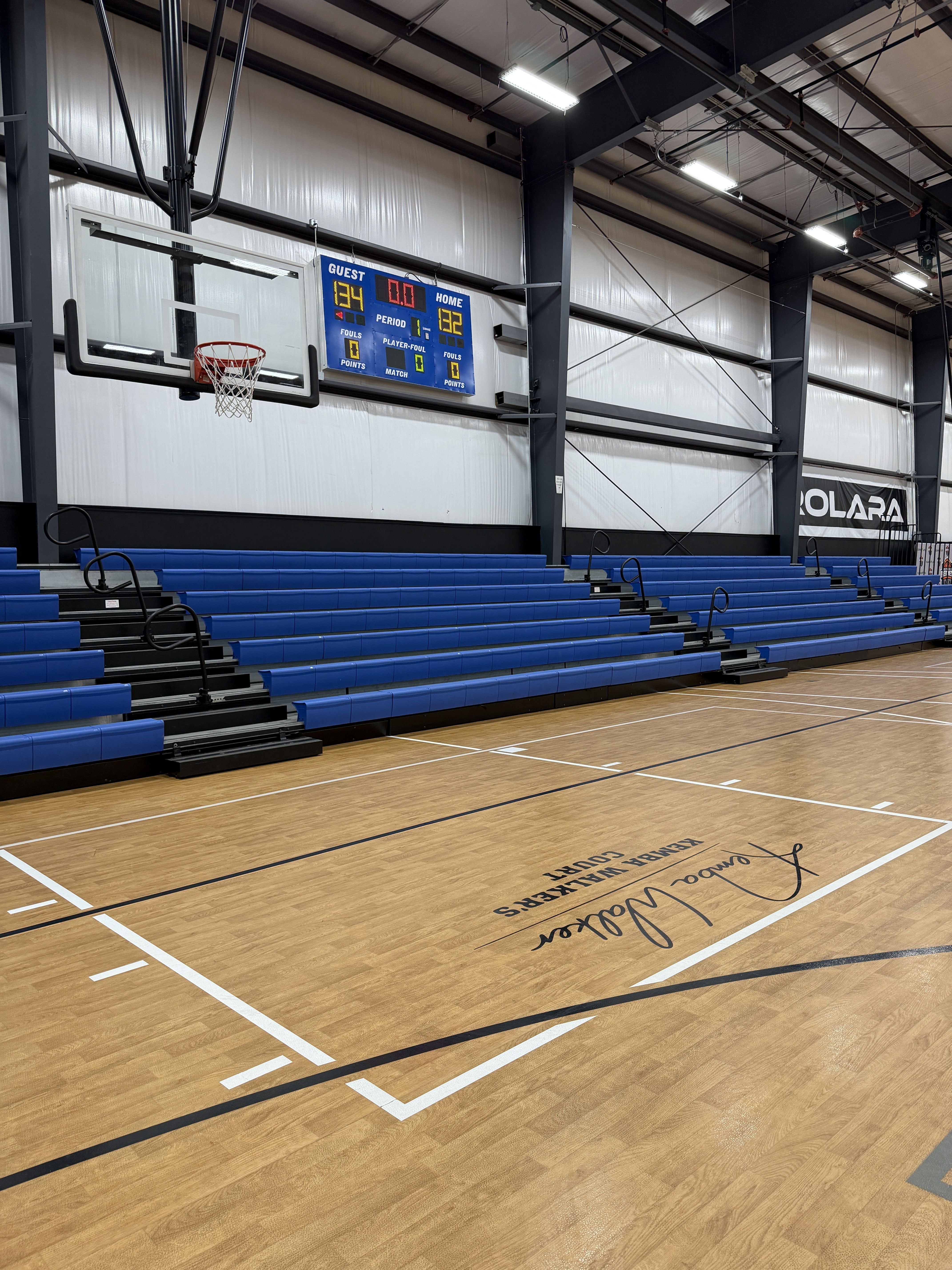 Indoor basketball gym with blue bleachers on the left, a hanging hoop and net, and a blue scoreboard showing scores and period. Steel beams, bright overhead lights, and a wooden court.