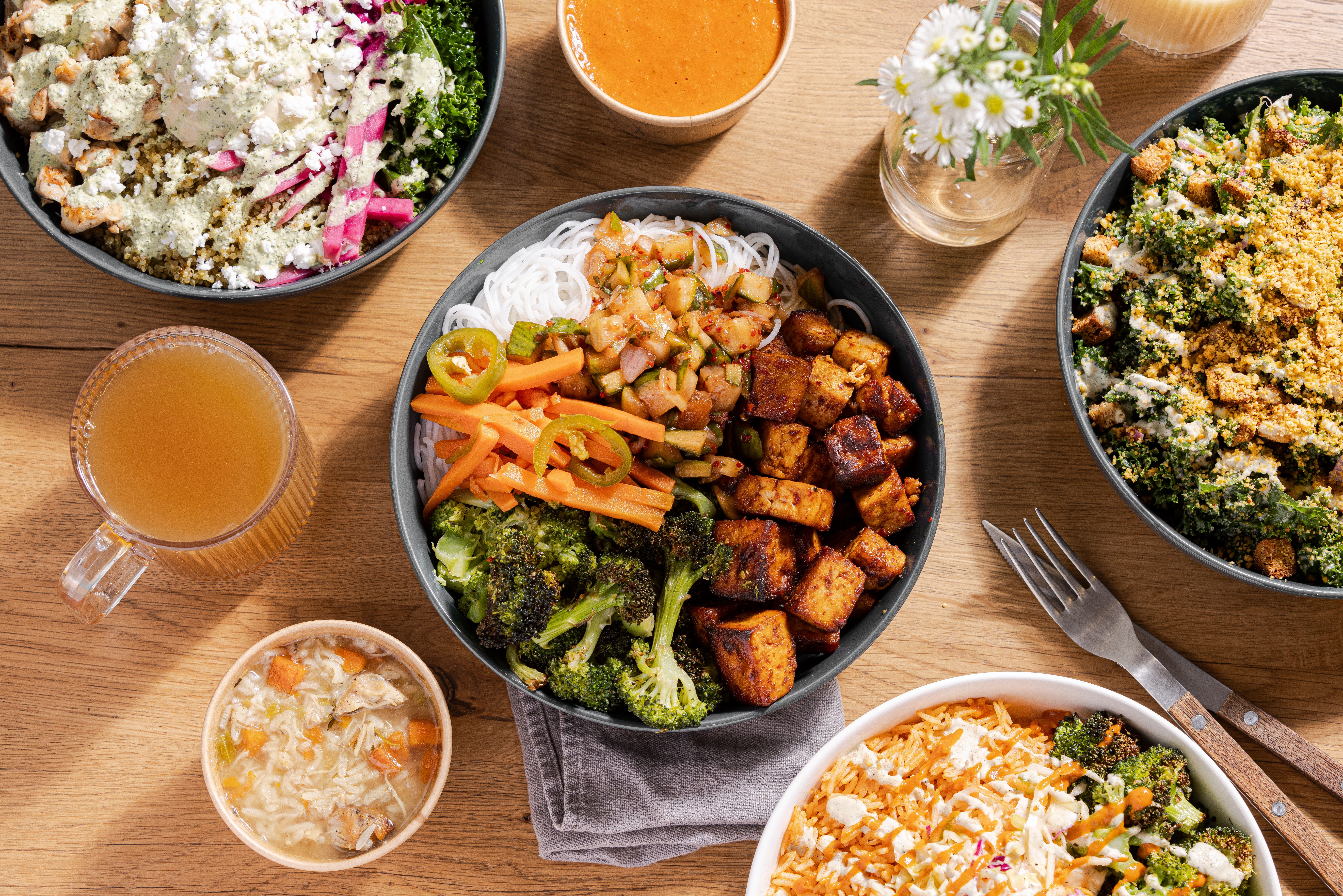 Top-down view of five bowls with various colorful foods including grilled tofu, broccoli, rice, pickled vegetables, kale salad with dressing, and sides like soup and a light brown drink on a wooden table.