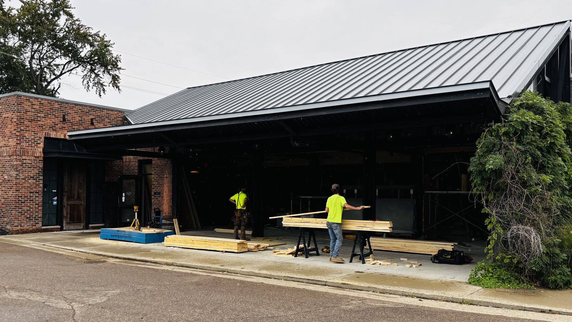 Brick building with a large gray metal roof over an open storefront under construction. Two workers in neon yellow shirts stand among stacked lumber on sawhorses; a blue toolbox sits nearby.