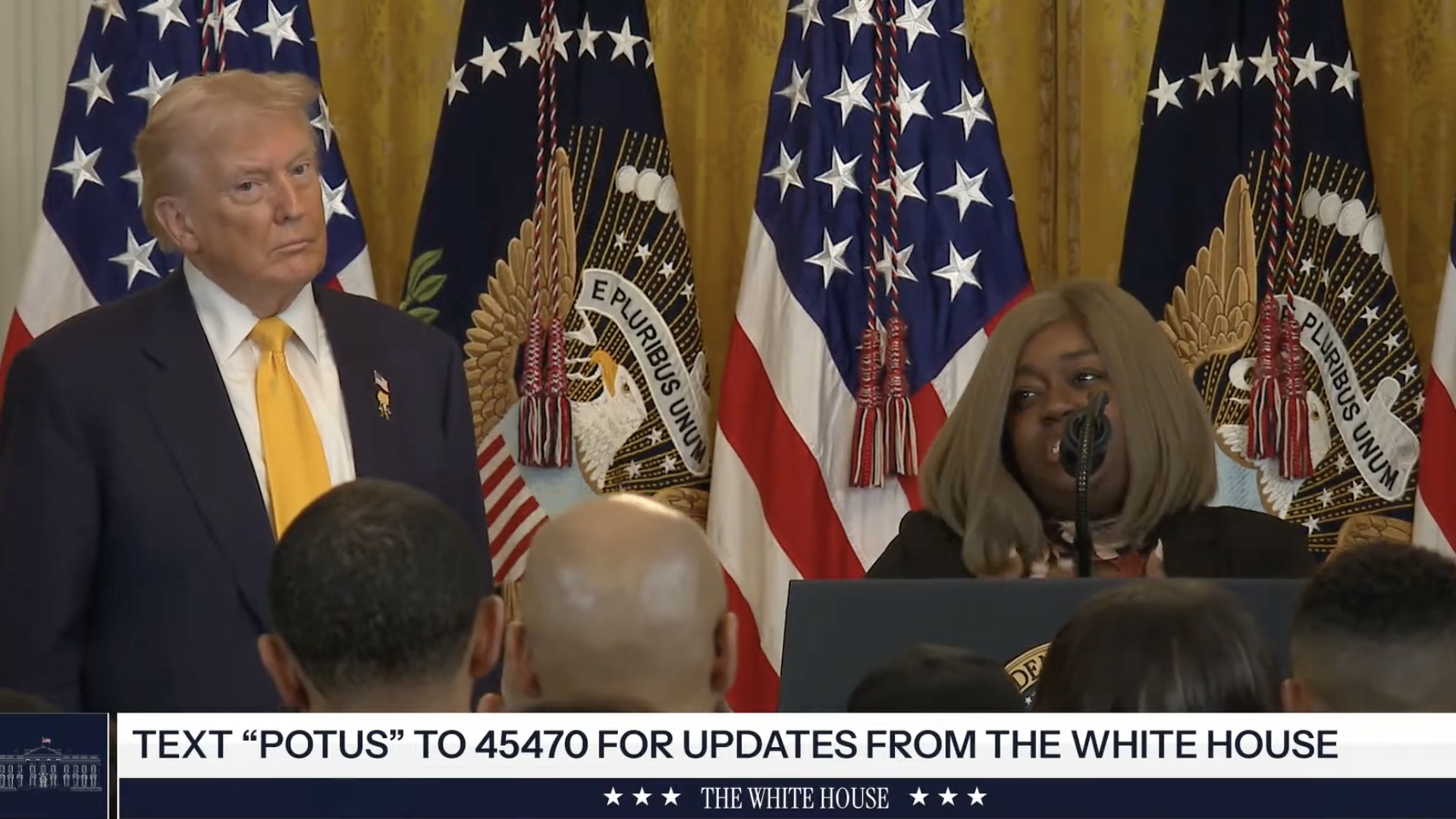 President Donald Trump stands beside a woman speaking at a White House podium, with U.S. flags behind them.
