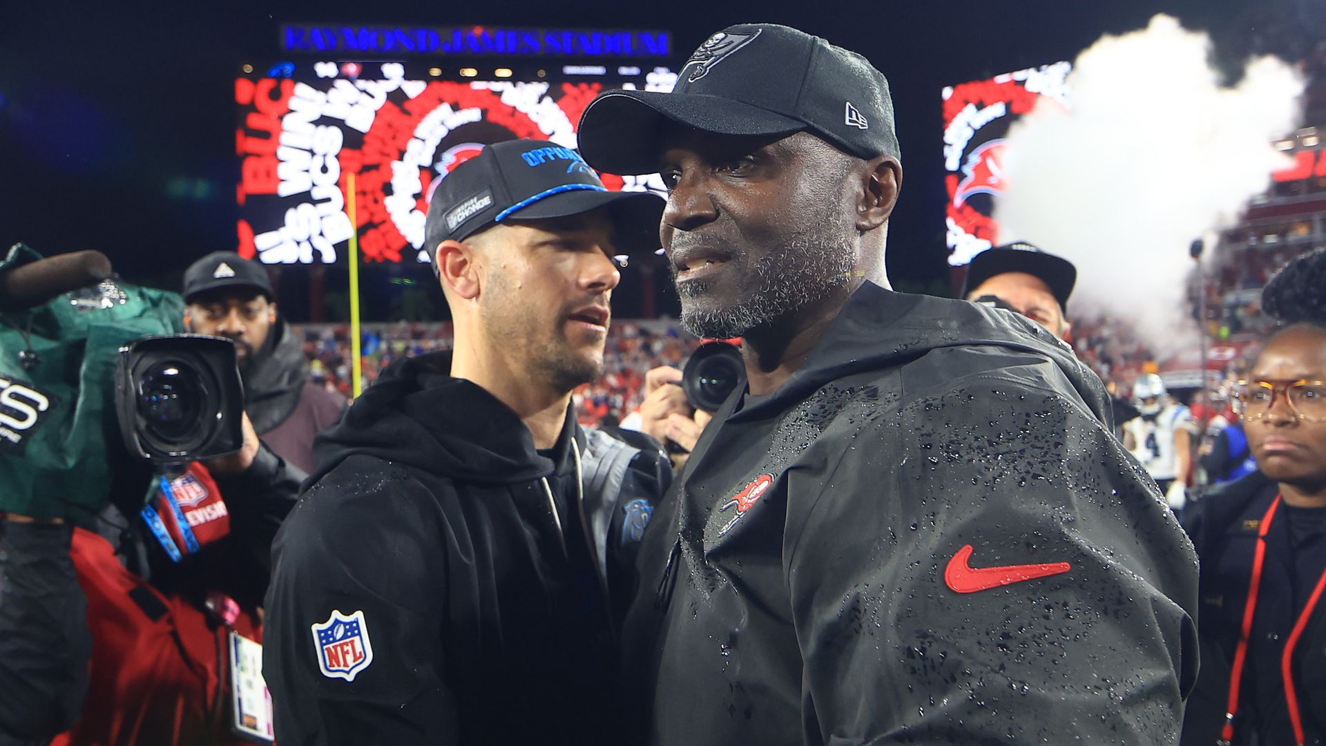 Buccaneers head coach Todd Bowles, a Black man with gray stubble in black athletic gear and a black team hat, shakes hands with Carolina Panthers coach Dave Canales after Tampa Bay's season-ending win.