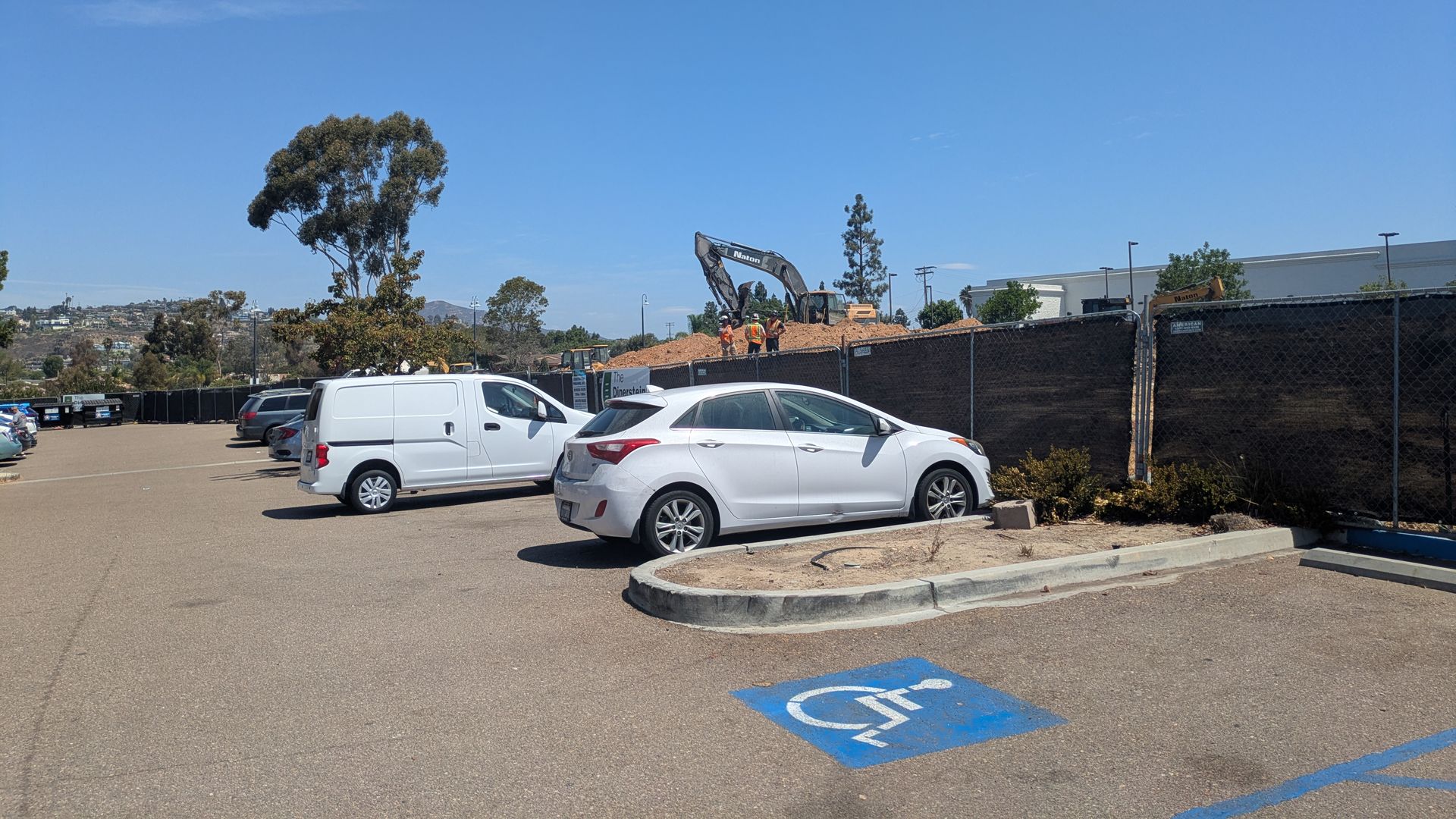 Blue sky over a parking lot with white vans; a chain-link fence and construction site with excavators in the distance, and a wheelchair-accessible parking space marking in the foreground.