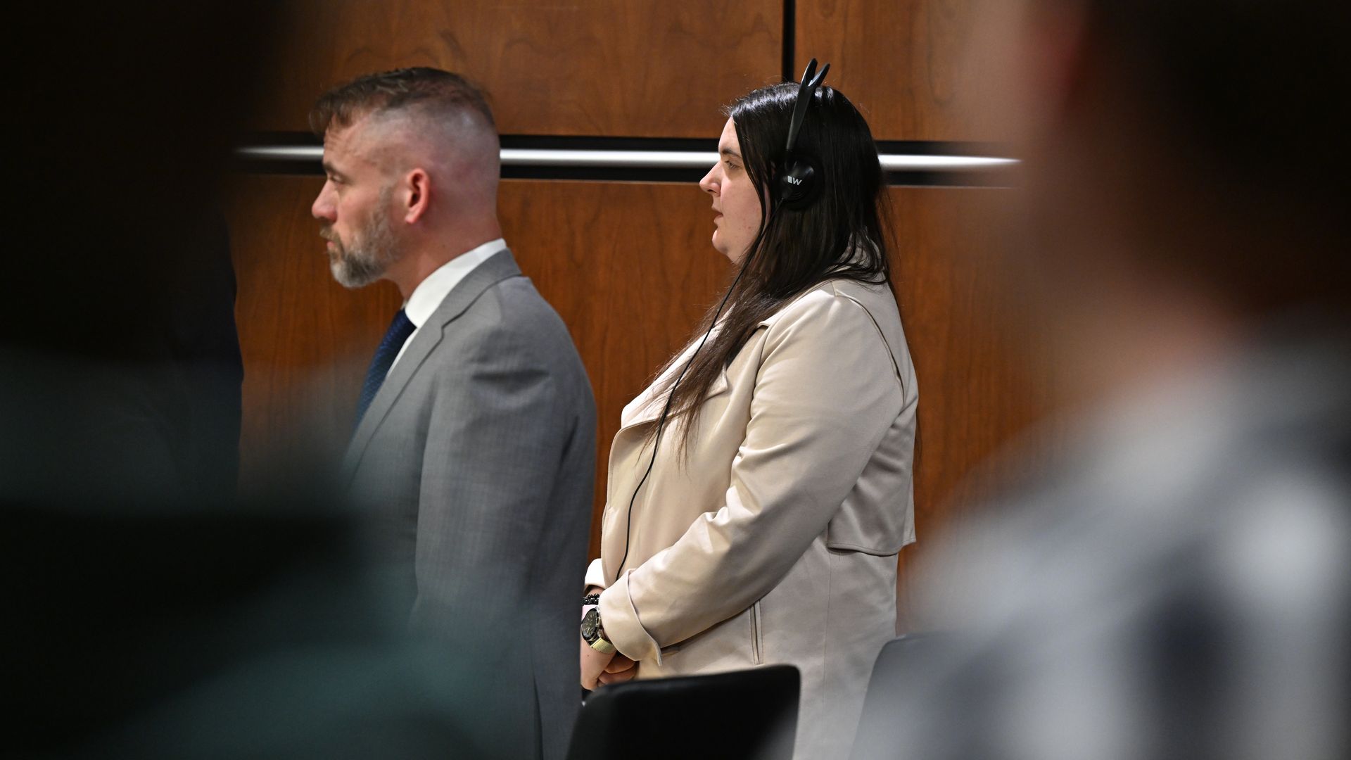 Side view of two people standing in a wood-paneled room; a defense attorney and Yeva Smilianska wearing a beige jacket and headphones.