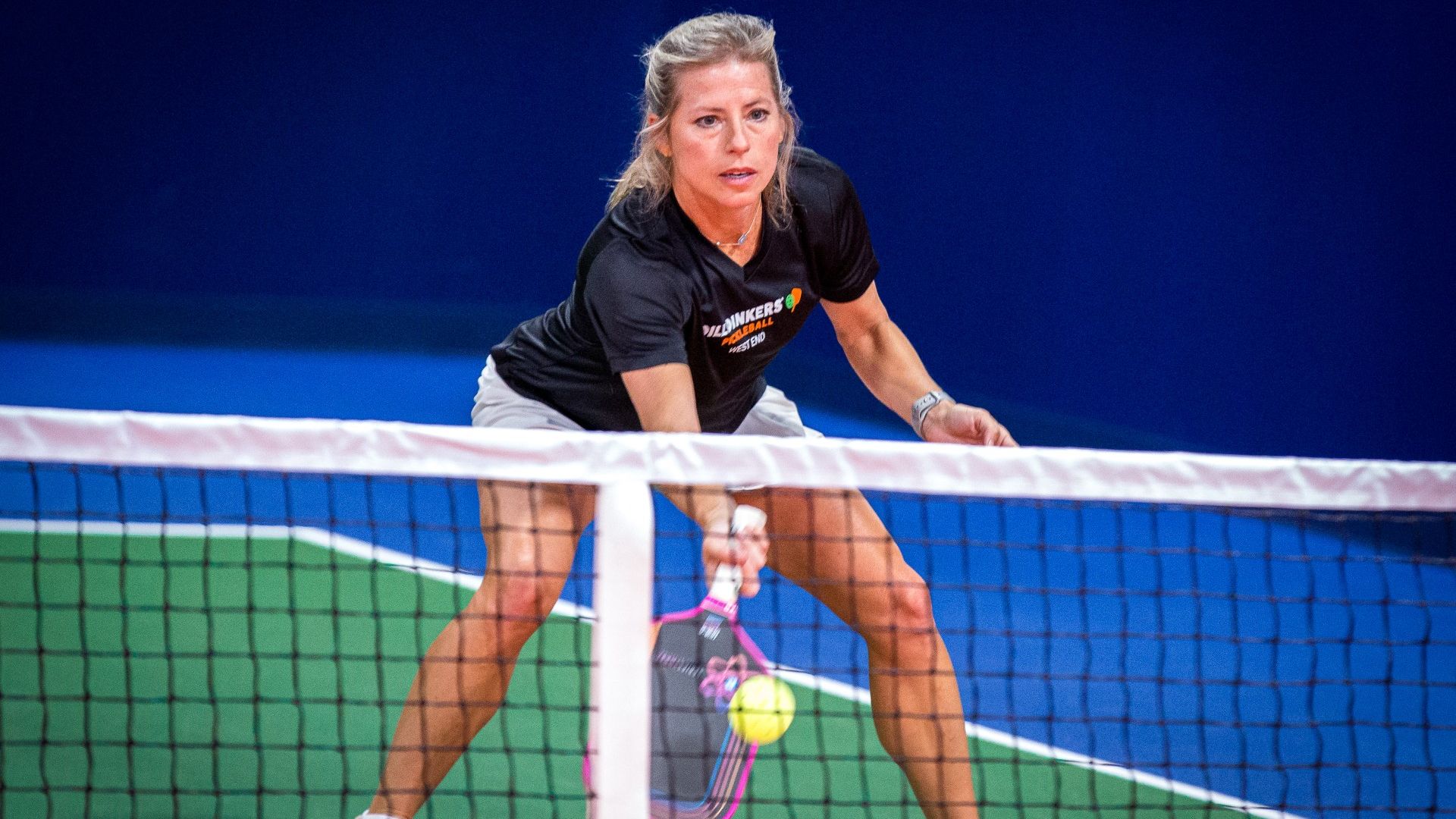 Female pickleball player in a black shirt and white shorts crouches at the net on a blue indoor court, ready to volley with a pink racket; net and green court visible.