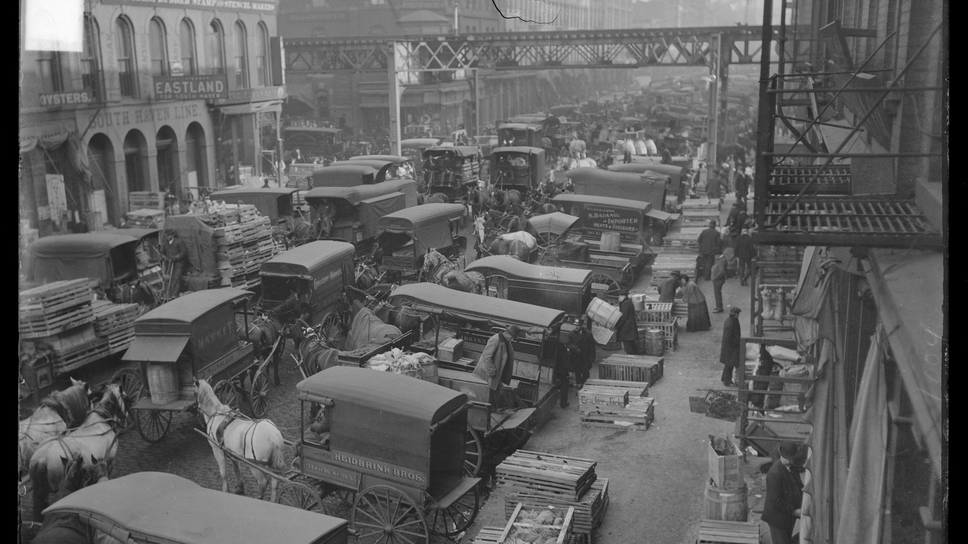 Black-and-white early 20th-century city market: crowded cobbled street with horse-drawn wagons, early trucks, stacked crates, and vendors beside storefronts under a metal bridge.