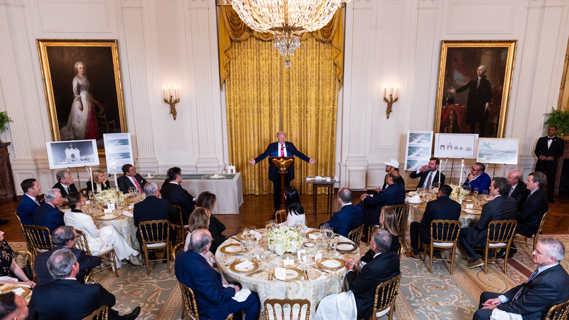 川普站在講台上講話，他張開雙臂，房間裡有幾個人圍坐在圓桌旁。