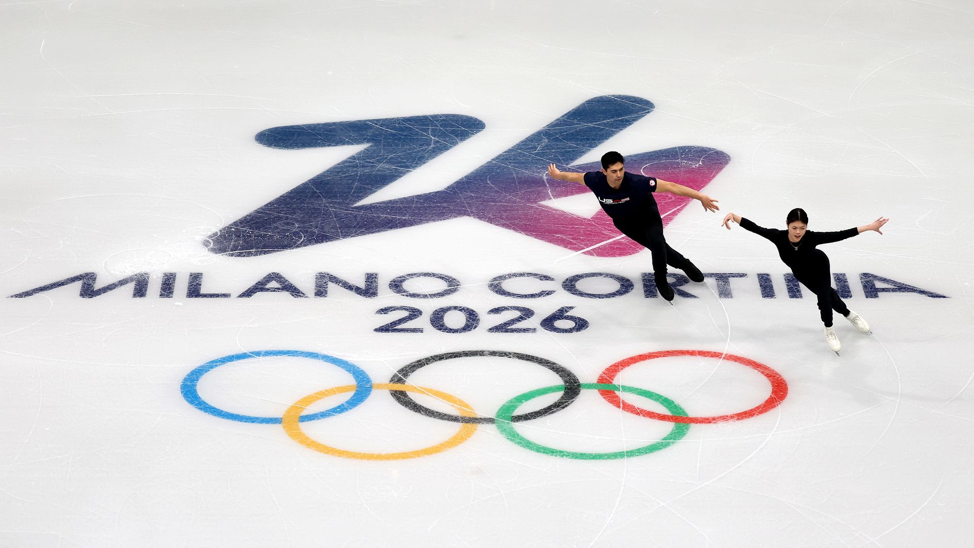 Two figure skaters in black outfits skating on ice with the Milano Cortina 2026 logo and Olympic rings visible beneath them.