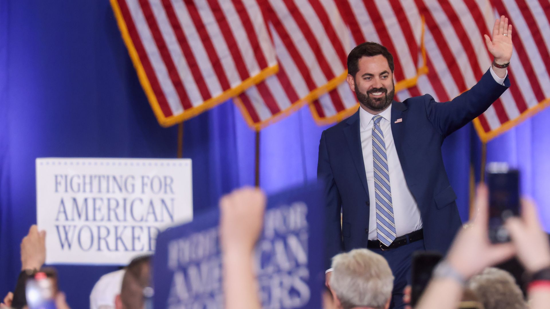 A man in a blue suit and striped tie waves to a cheering crowd at a political rally, with American flags in the background and signs reading "Fighting for American Worker".