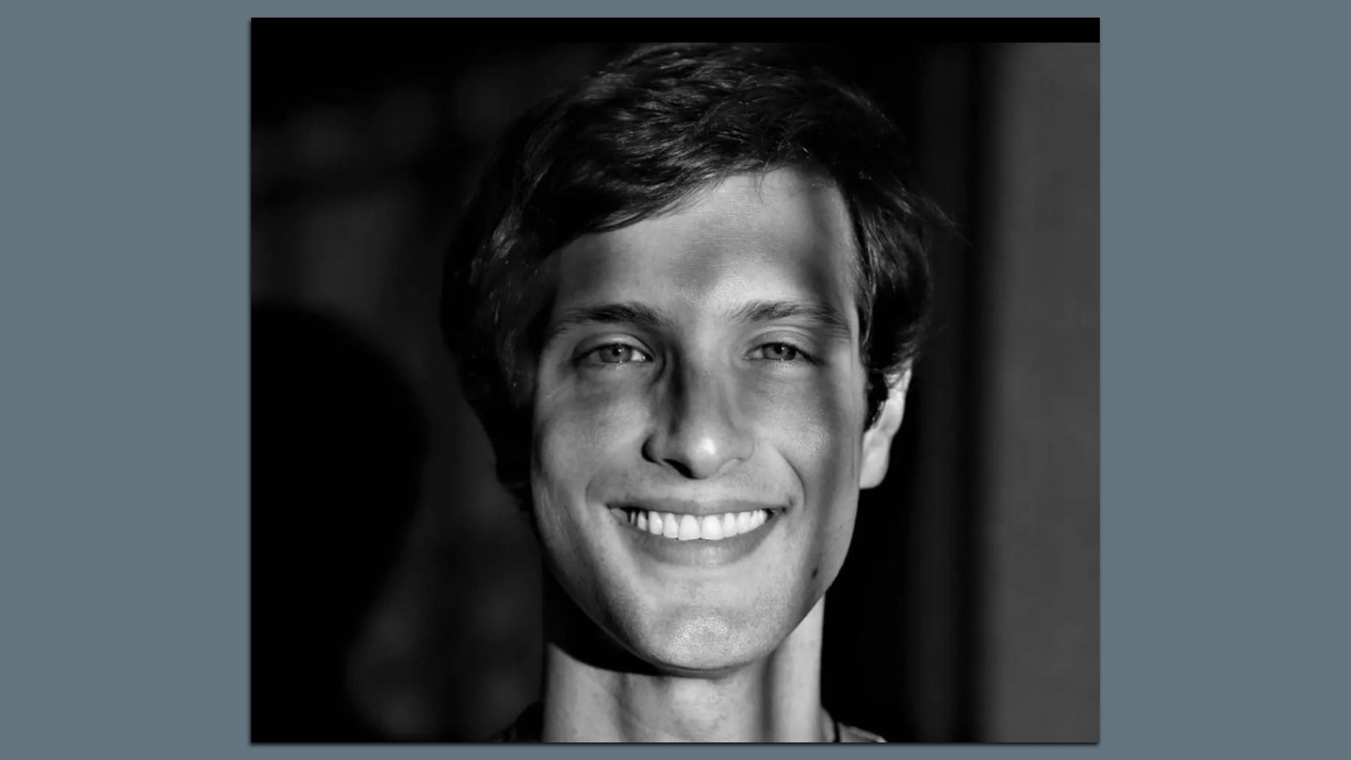 Black-and-white close-up portrait of a smiling young man with short dark hair, visible teeth, and a dark, blurred background.