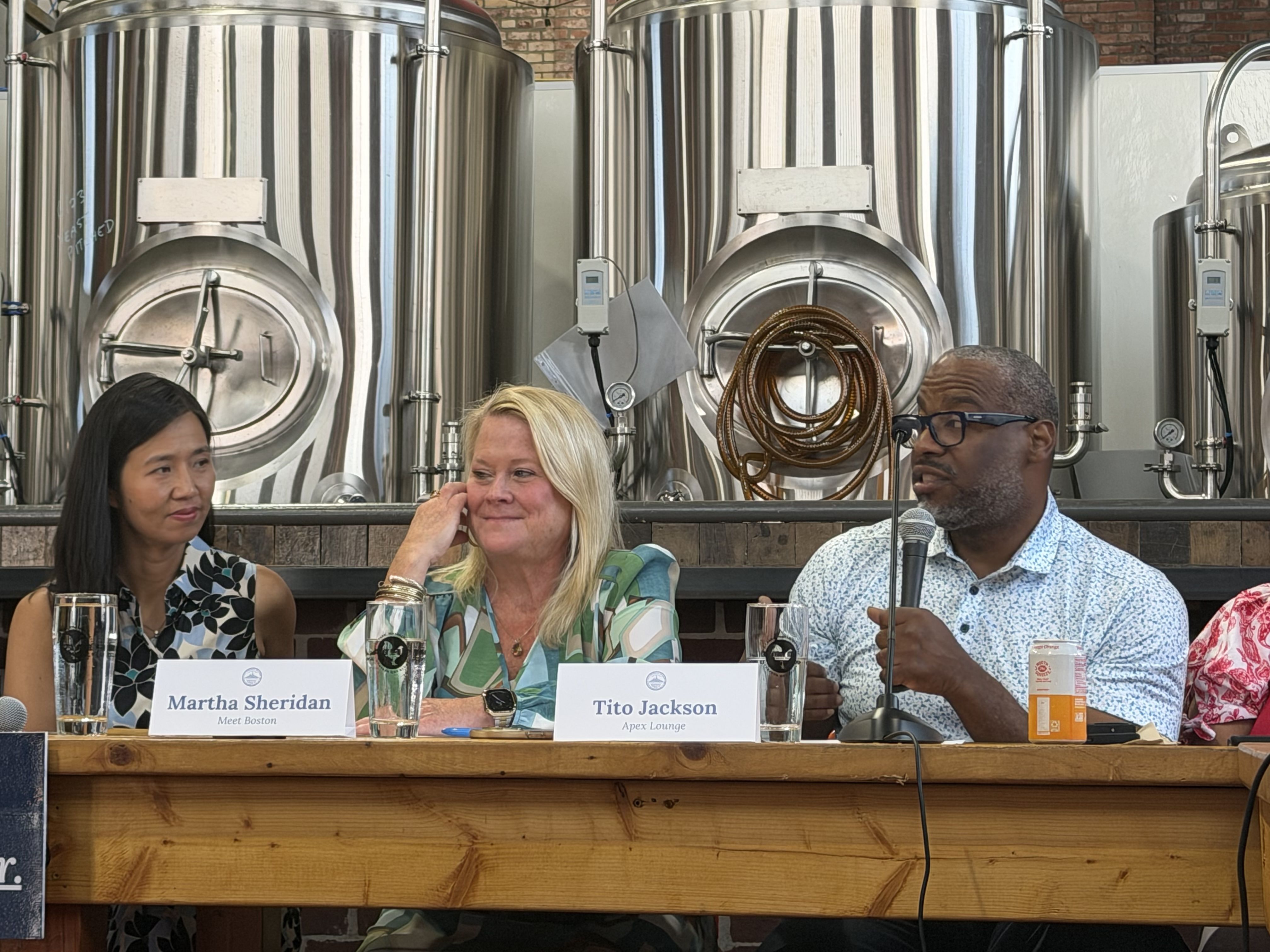 A close-up shot of three Boston leaders speaking at a roundtable at Roundhead Brewing in Hyde Park: Tito Jackson, right, holds the mic and discusses the local economy, next to Martha Sheridan, center, and Boston Mayor Michelle Wu, left.