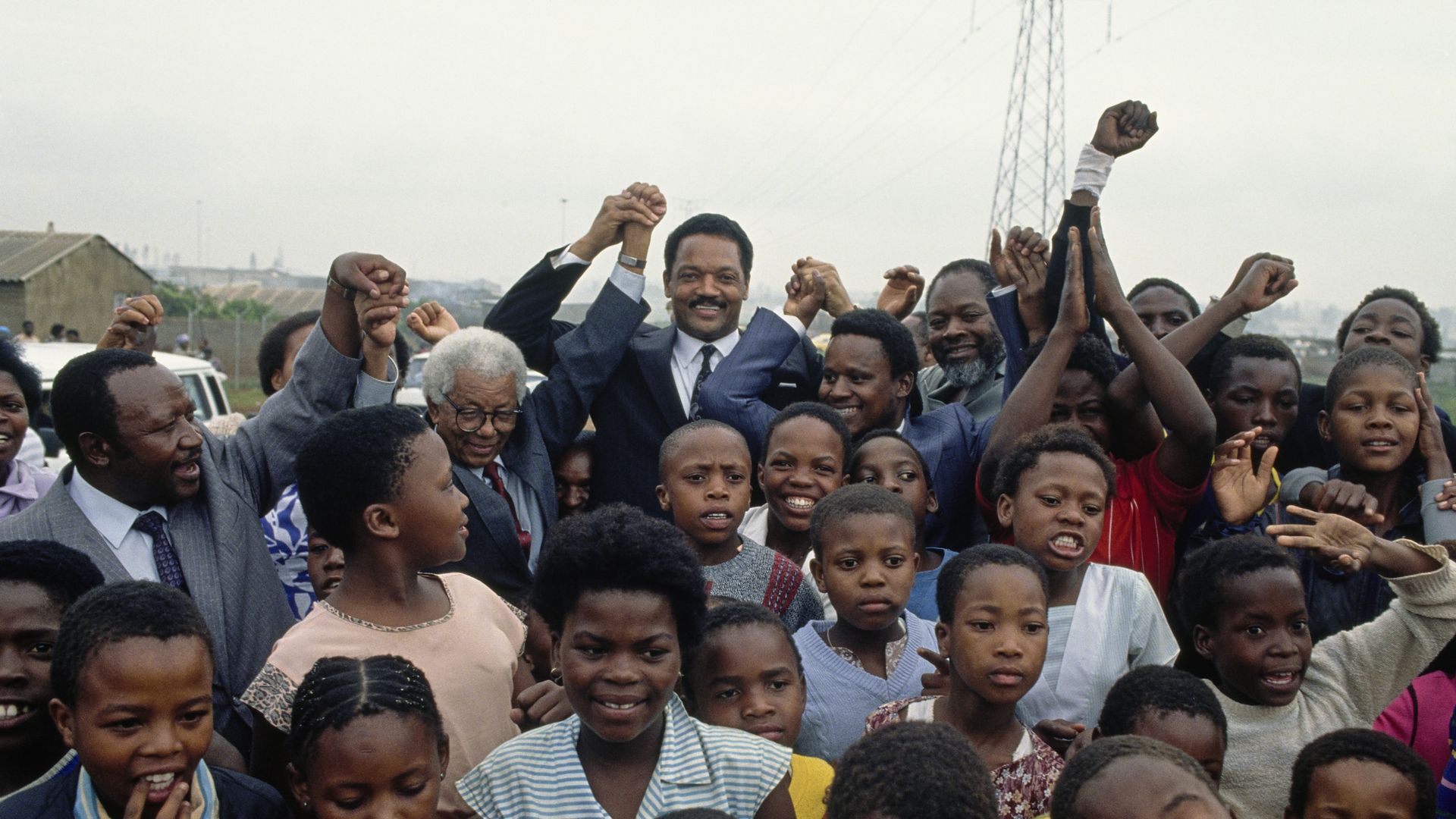 Group of smiling Black people in Soweto, Johannesburg, South Africa, including children and adults, some holding hands raised, outdoors near houses and power lines on a cloudy day with Jesse Jackson.