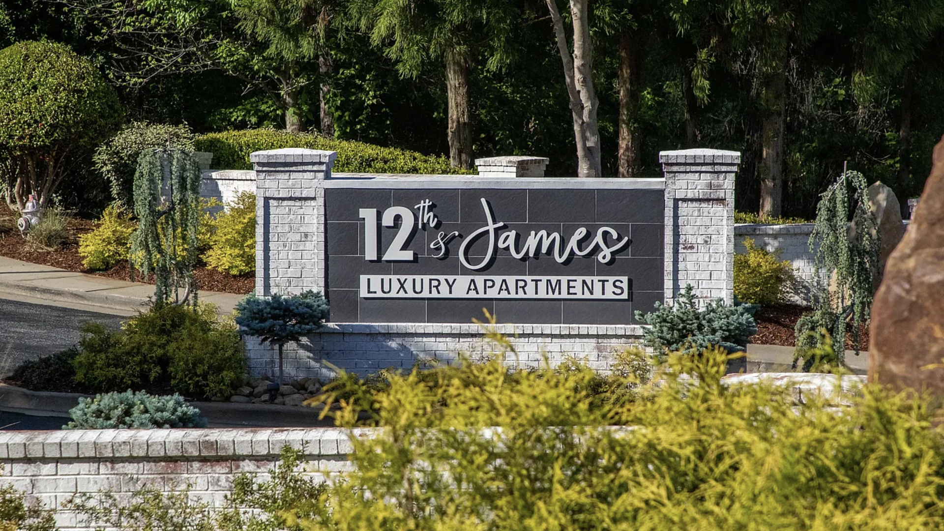 White brick sign with black background reading "12th & James Luxury Apartments" surrounded by green and yellow bushes and trees in Atlanta.