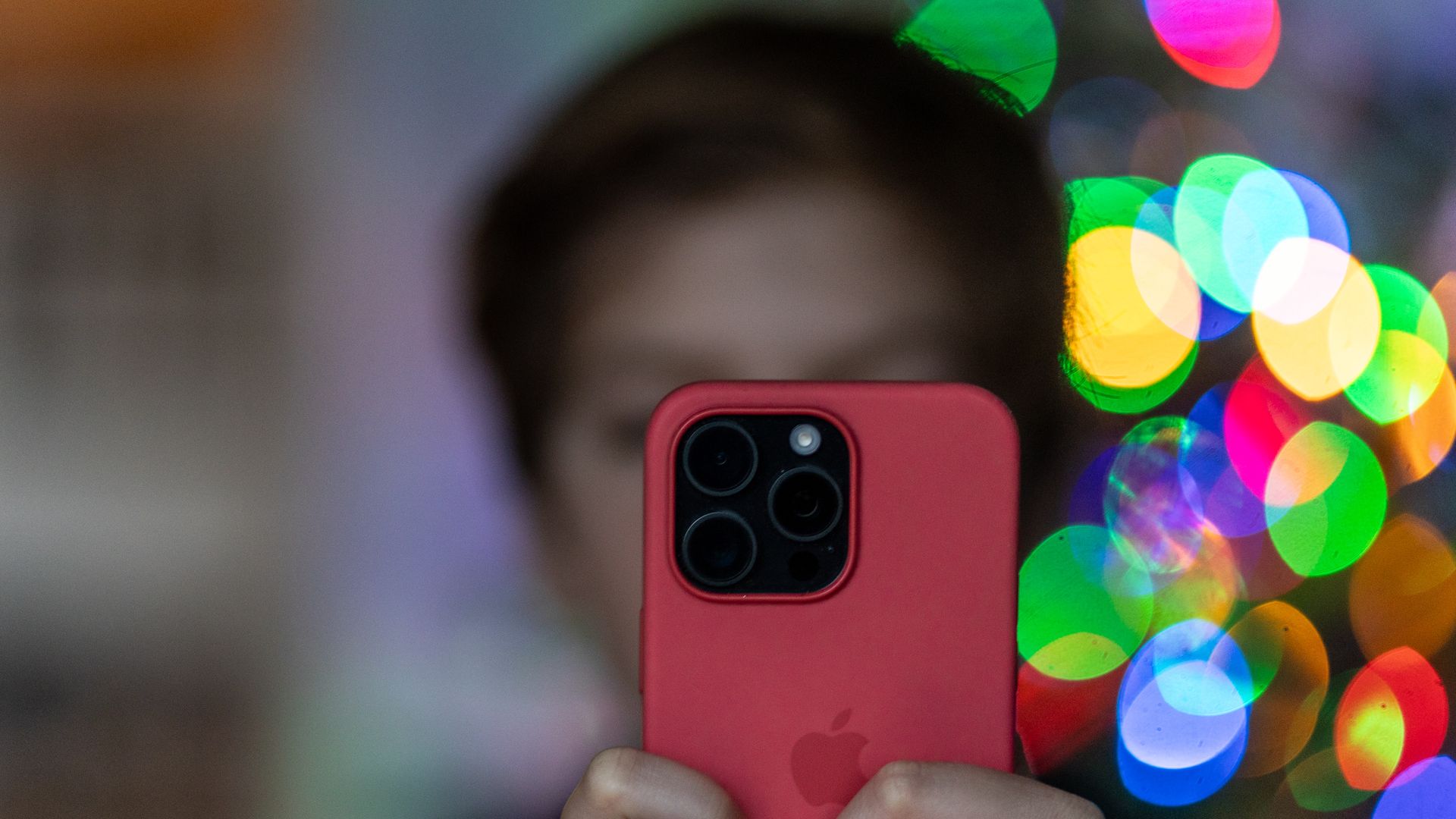 A child holds an iPhone in a red case. The background is blurred. 