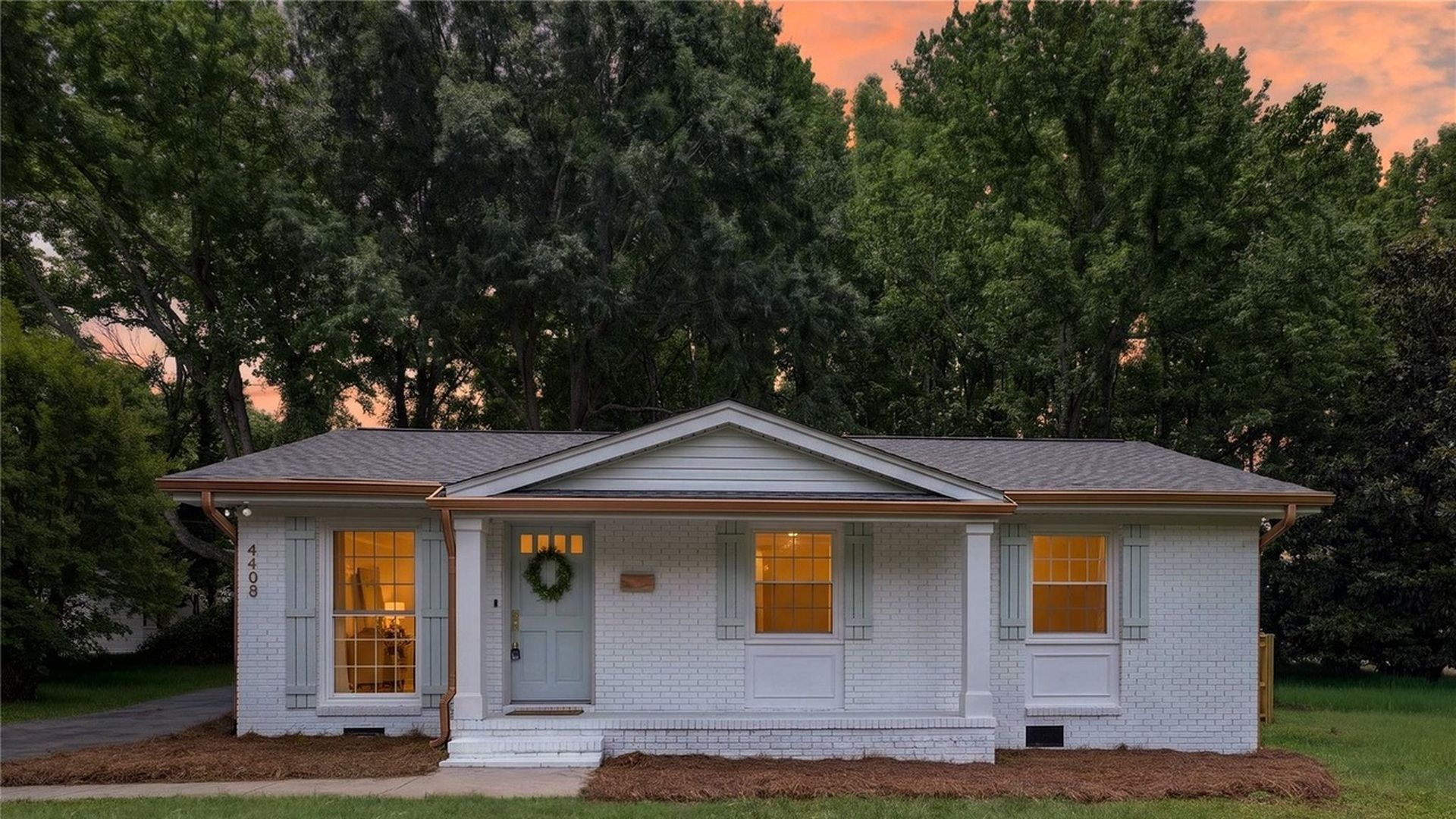 hot homes. White brick, single-story house with a light blue door and wreath, three lit windows with gray shutters; green lawn, mulched beds, orange sunset sky behind trees. For Sale watermark bottom right.