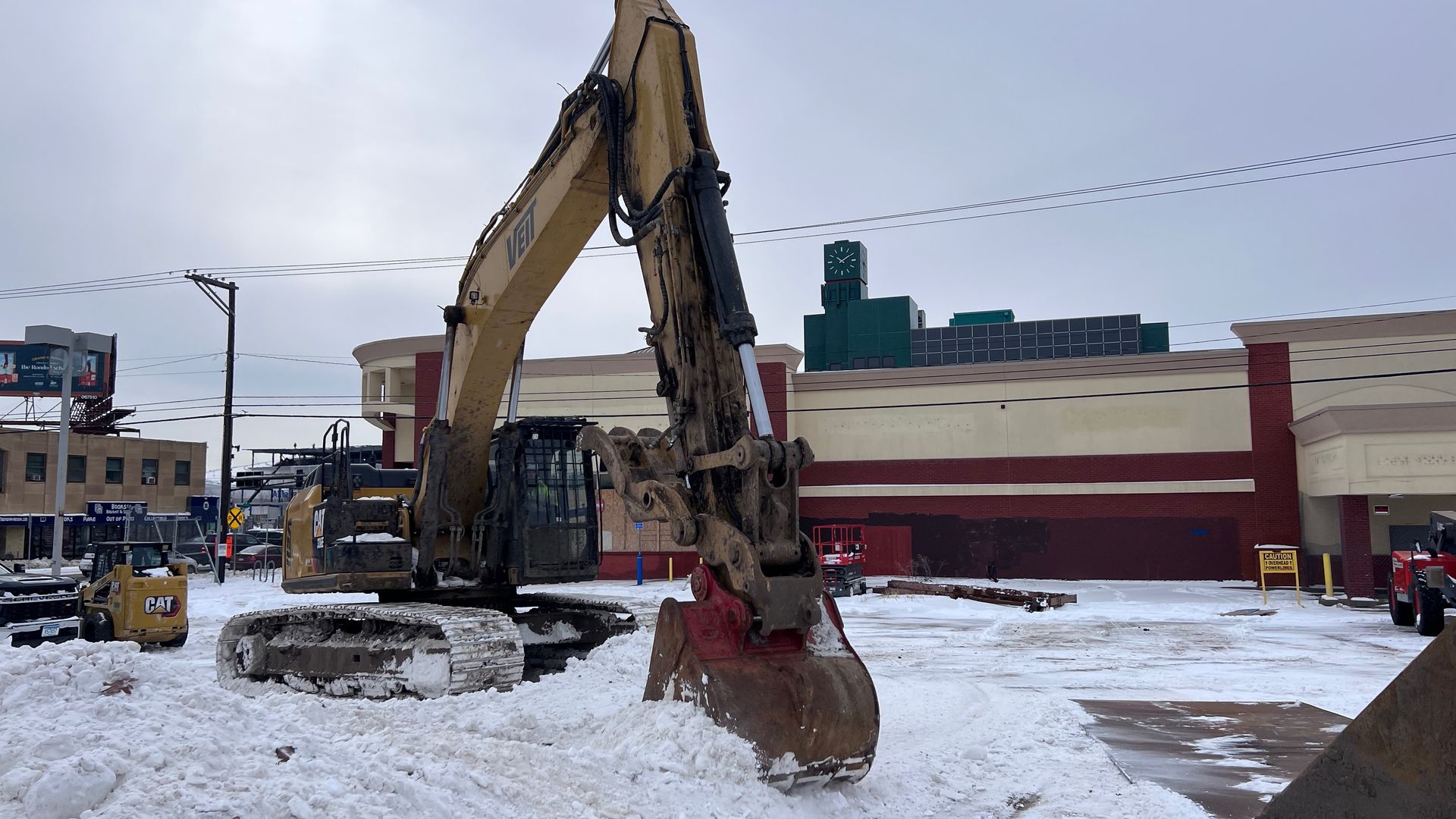 Winter construction site with a large yellow excavator and red bucket in snow, adjacent to a shopping center with a green clock tower and power lines in the background.