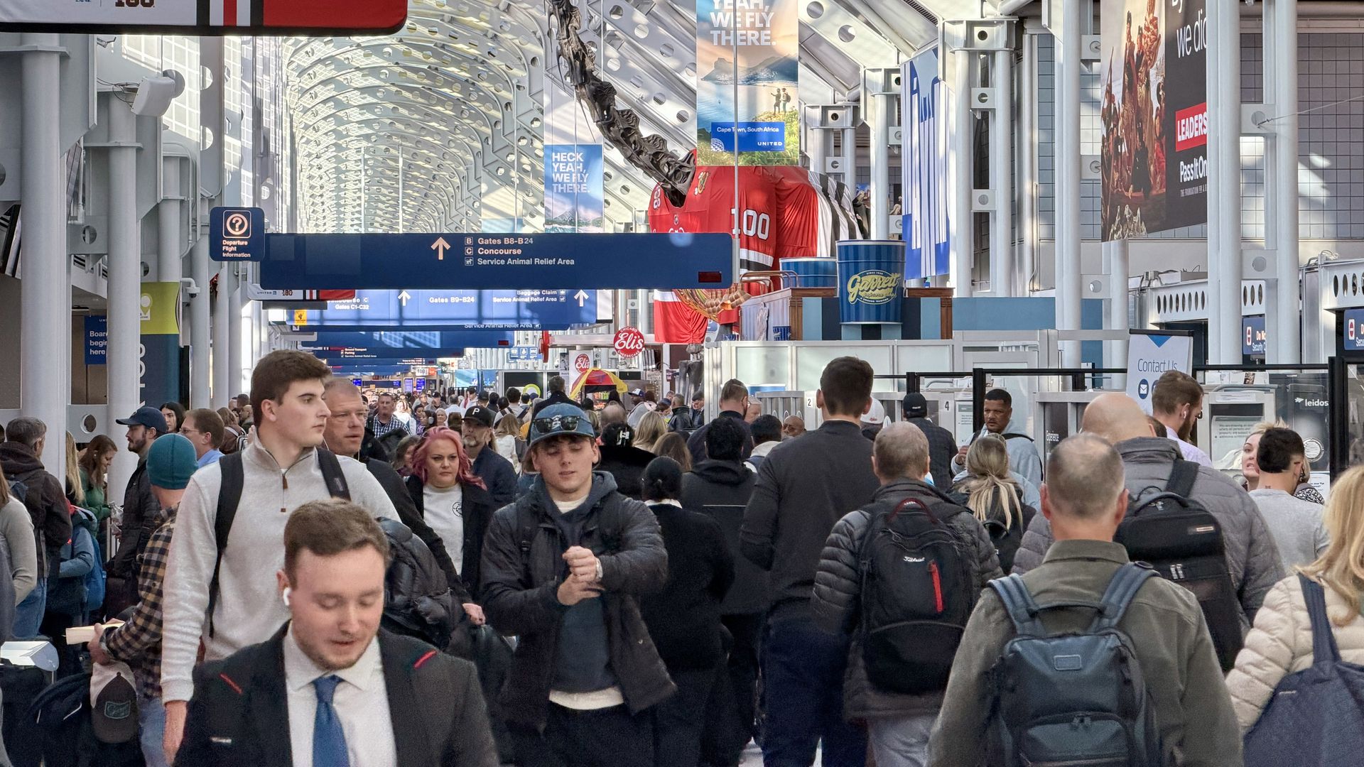 Crowded airport terminal with travelers walking, overhead signs for gates B7 and B8-B24, a large dinosaur skeleton, red sports jersey display, and bright natural light through glass ceiling.
