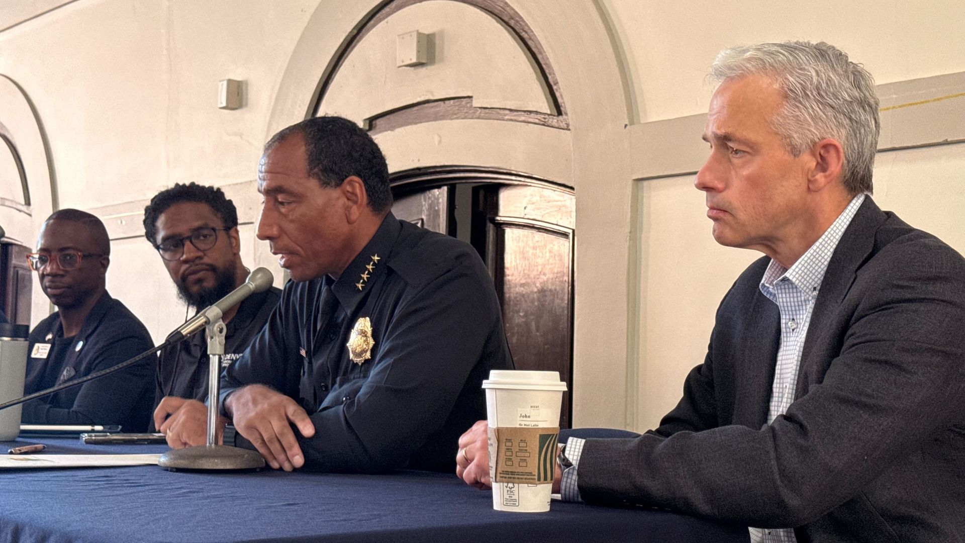 Panel of four men sits behind a blue table: a police officer in uniform speaking into a microphone, two Black men in dark shirts with glasses, and a gray-haired man in a suit listening; a coffee cup sits at the right.